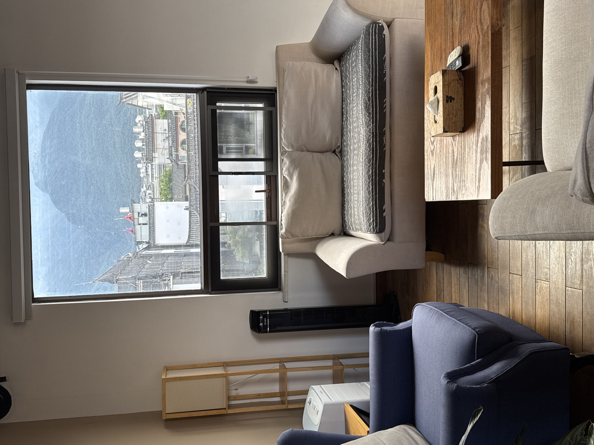 This is an interior shot of a living space with a large window offering a view of Dali City, China. The room features a blue armchair on the left, a light-colored loveseat in the center with a patterned throw on its seat, and a tan armchair partially visible in the foreground. A wooden coffee table sits in front of the loveseat, holding a wooden tissue box and two remote controls. To the left of the loveseat, a tall black tower fan stands next to a wooden shelf unit containing a white air purifier. The window looks out onto a city skyline with traditional tiled roofs and mountains in the background. The light suggests it is daytime.