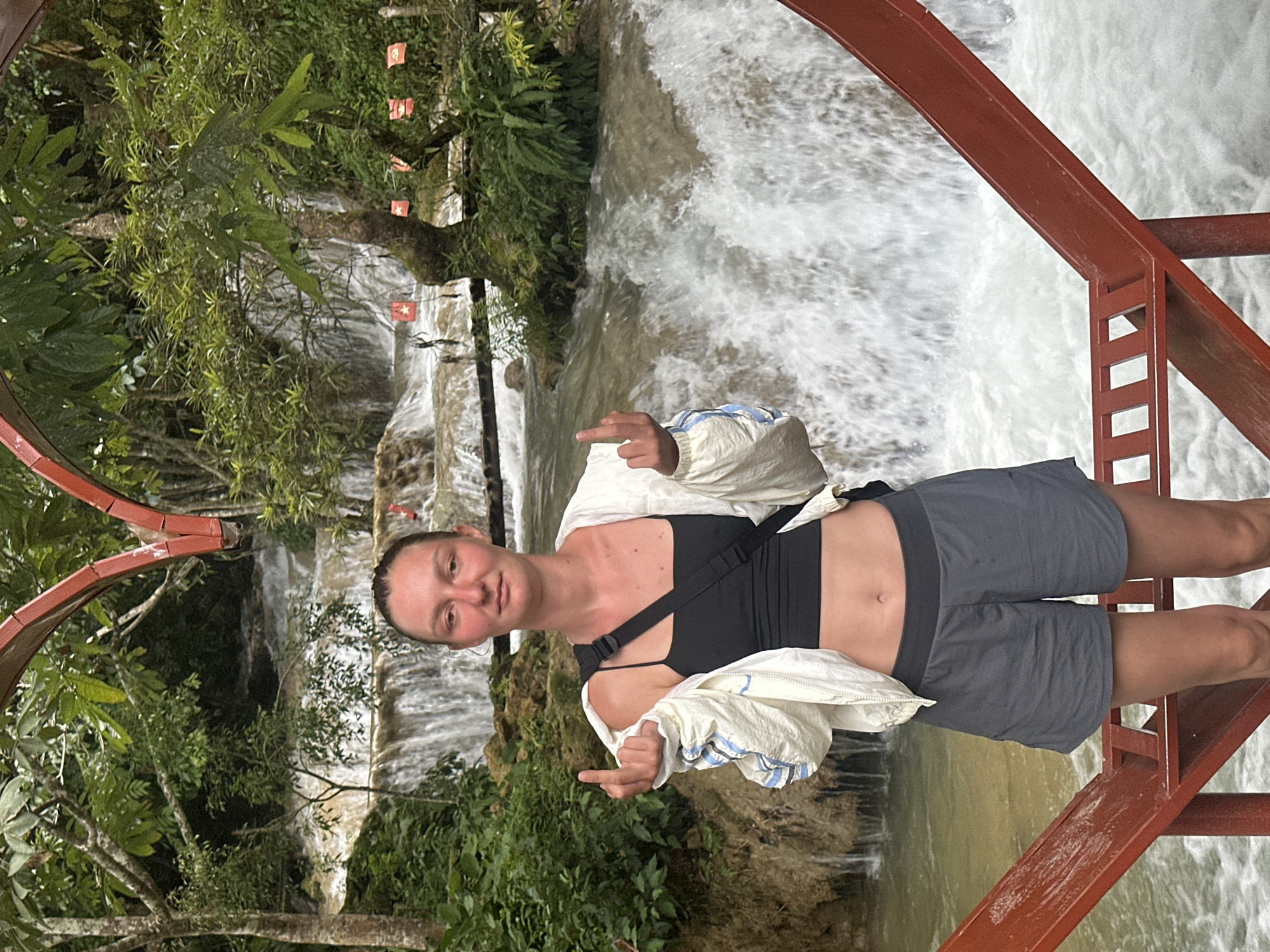 A woman stands on a red metal platform in front of a waterfall in a lush, green environment. She is wearing a black tank top, a white jacket with blue and white striped accents, and grey shorts. Her hair is pulled back in a bun. She is giving a middle finger gesture with both hands. Small red flags with white stars are visible on a tree branch behind her. The waterfall appears to be tiered, with water cascading down multiple levels. The overall scene suggests a tourist location. The location in the city of Cổ Lũng Commune, Vietnam is not evident from the image itself, but the context of lush vegetation and tiered waterfalls is consistent with such locations.