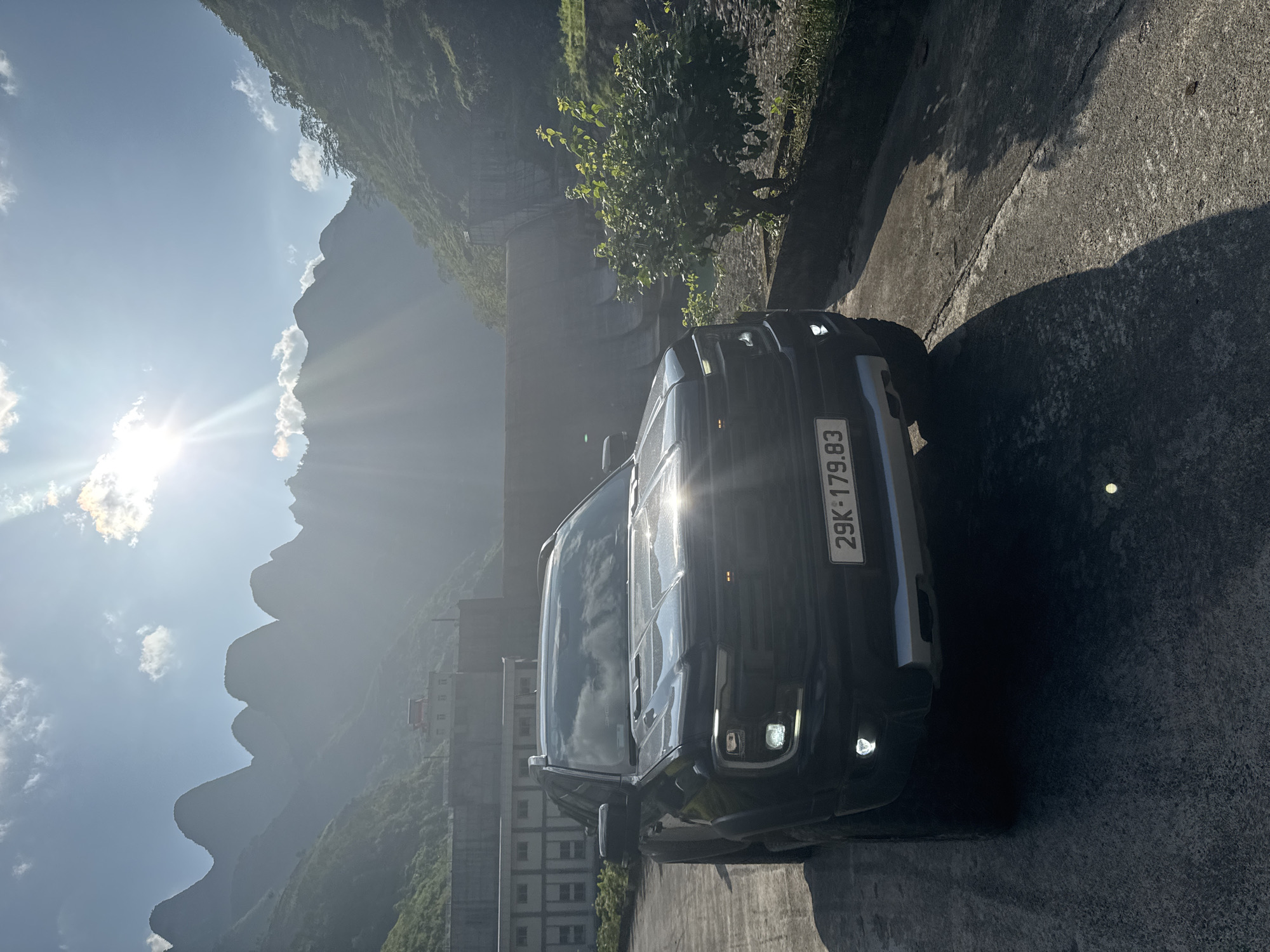 A black SUV is parked on a paved surface, likely a road or parking area. The car has a license plate that reads "29K-179.83." The sun glares off the vehicle's hood. Behind the car is a mountainous landscape partially obscured by haze. Buildings, including what appears to be a multi-story structure, are built into the mountainside. A tree grows along the paved area next to the car. This image depicts a scene within the Mèo Vạc Commune, Vietnam.