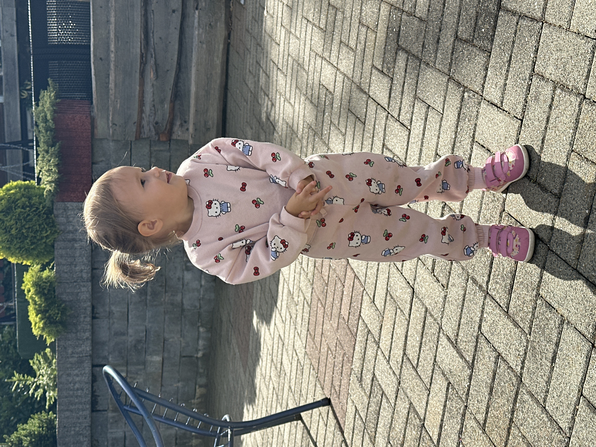 A young child, appearing to be a toddler, stands outdoors on a paved patio area. The child is wearing a pink pajama set with a Hello Kitty and cherry print. They have their hands clasped in front of them and are looking to the right. The background features a stone wall, some greenery, and a blue metal object, possibly a piece of outdoor furniture. The lighting suggests it is daytime, with shadows indicating sunlight. The location is identified as Wisła, Poland.