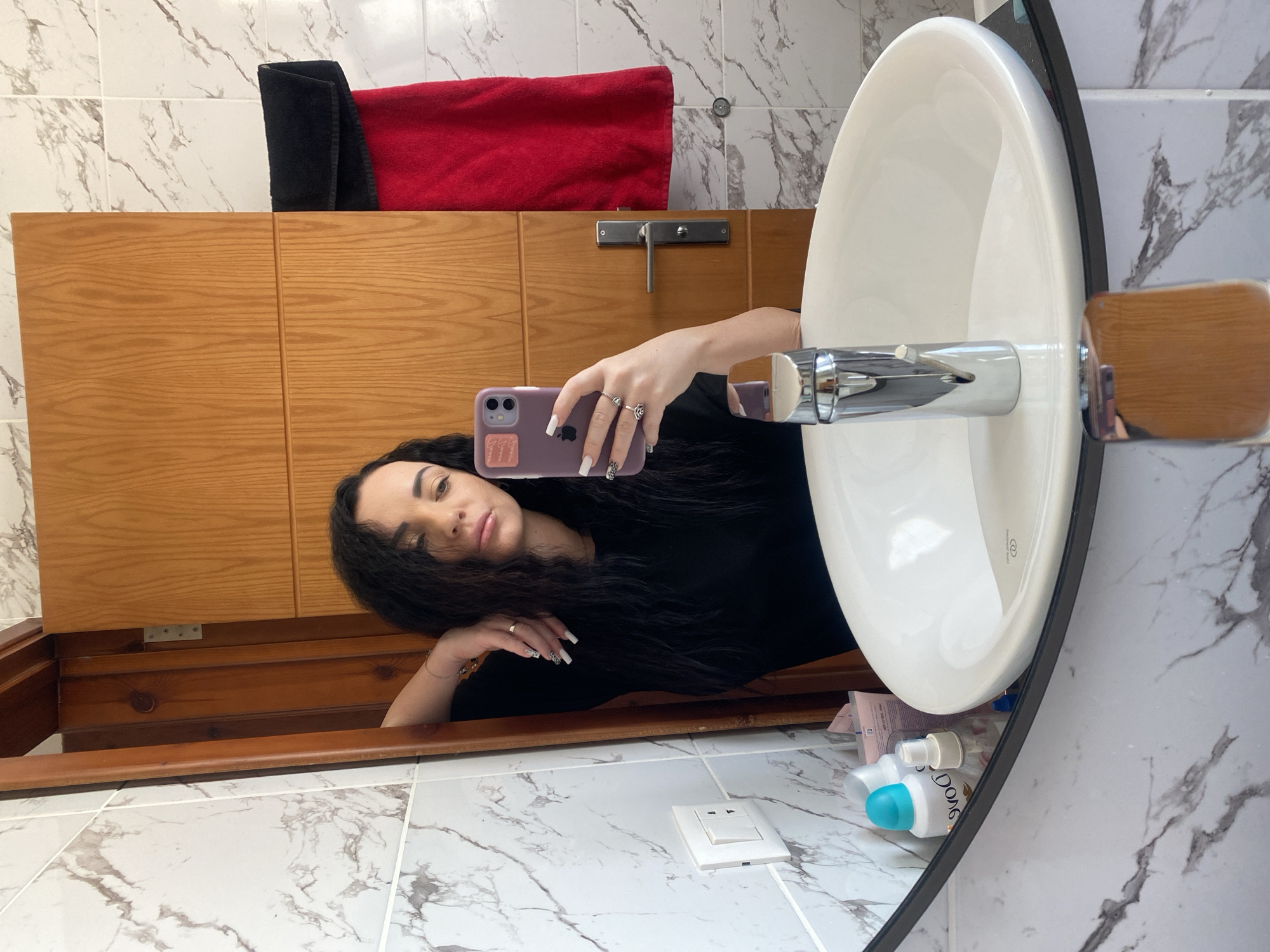 In Sharm El Sheikh, Egypt, a woman with long, dark, wavy hair takes a mirror selfie in a bathroom. She is wearing a black t-shirt and multiple rings on her fingers. Her left arm is resting on a wooden door frame. In front of her, a white sink with a chrome faucet is visible, along with various toiletries on a shelf. A red towel and a black towel are hanging on the wooden door. The bathroom walls are tiled with a marble pattern.