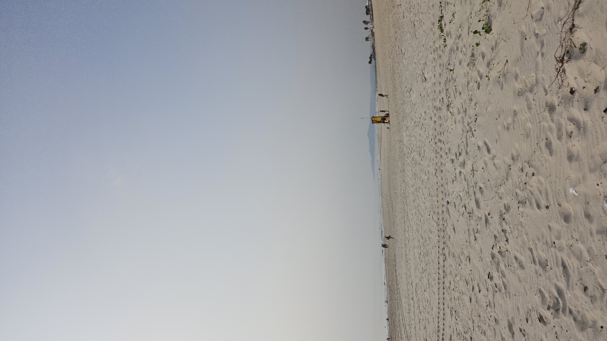 The image displays a vast, tranquil beach scene under a clear, bright sky, likely in Bac Cam Ranh, Vietnam. The horizon line runs horizontally across the middle of the frame, separating the expansive sky from the wide sandy beach.
In the mid-ground, a prominent yellow and red lifeguard stand is situated on the sand. A few small, indistinct figures, presumably people, are visible on the beach; one appears near the lifeguard stand, while others are further along the shore towards the water's edge, seemingly walking or standing. The sand itself is a light tan color, covered with numerous footprints and gentle undulations. Towards the foreground and right side of the image, sparse beach vegetation, including green vines and dried twigs, can be seen on the sand.
The sky above is a uniform light blue to hazy gray, suggesting a sunny day with possibly some atmospheric haze. In the far background, a faint line of distant mountains or hills is visible along the horizon. The ocean appears as a thin strip of water at the very edge of the beach.
The overall atmosphere is one of serene openness and quiet. The light quality indicates it is daytime, possibly morning or late afternoon, given the soft shadows. No discernible text is visible anywhere in the image.