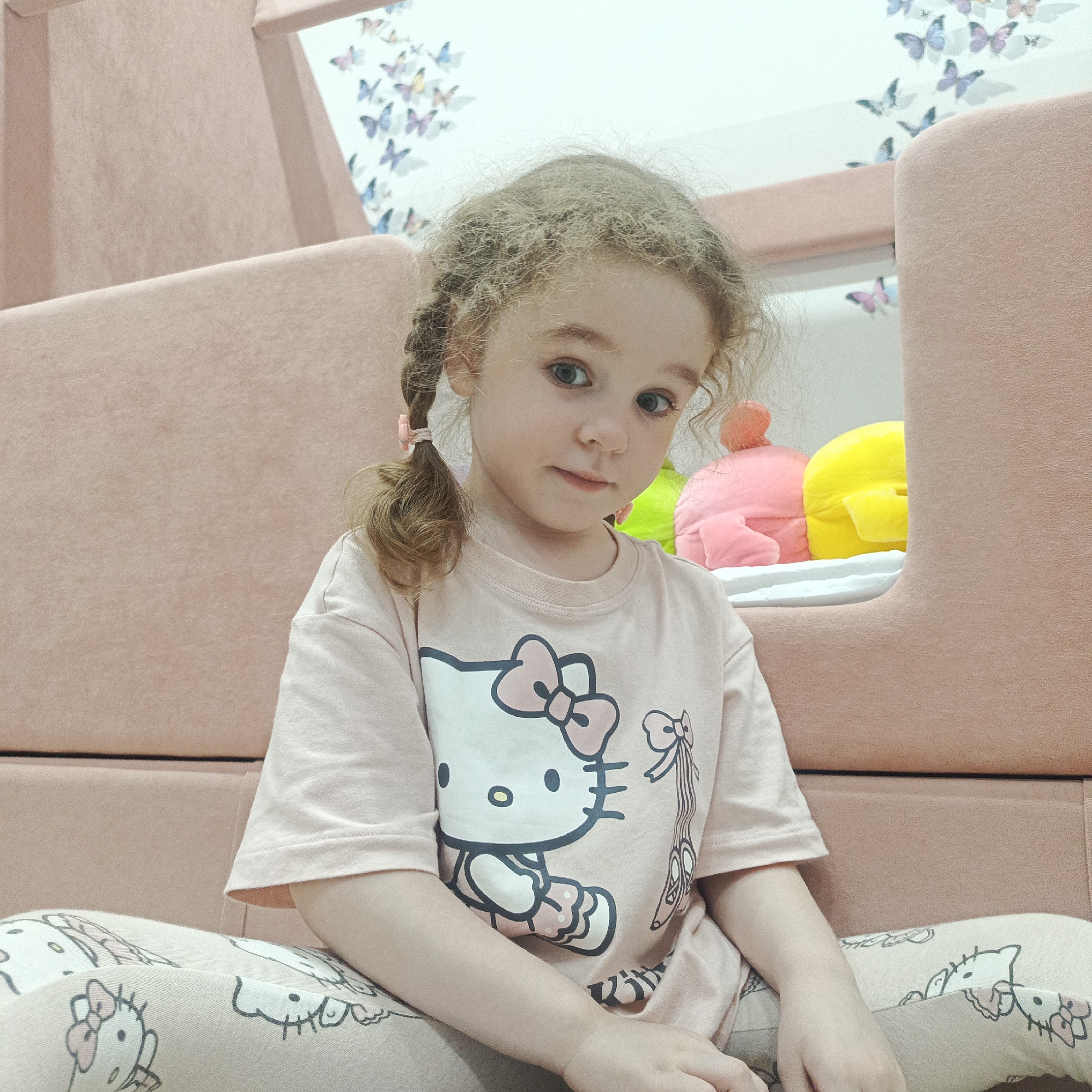 This media file features a young girl, likely between three and five years old, seated indoors and looking directly at the camera. She has curly, light-colored hair styled into two small pigtails fastened with pink ties, and her eyes are blue-grey.
The girl is wearing a light pink short-sleeved t-shirt adorned with a prominent illustration of Hello Kitty, complete with a pink bow. Her attire is completed by matching light pink pajama pants patterned with repeated Hello Kitty faces and bows. A small illustration resembling ballet shoes is also visible on the right side of her shirt.
The setting appears to be a child's room or play area, characterized by soft pink, upholstered modular furniture forming the background. Behind this furniture, a wall or headboard is decorated with numerous pastel butterfly decals in shades of pink, purple, and blue. To the girl's right, within a nook of the furniture, several brightly colored soft toys in pink, yellow, and green are visible.
The scene is brightly and evenly lit, suggesting daytime. The girl's expression is calm and direct, possibly shy or thoughtful. The overall ambiance is cheerful and child-friendly, reinforced by the decorations and toys. While the Hello Kitty brand is visually prominent, no specific text is explicitly readable. The provided location context is Markovskiy, Russia.