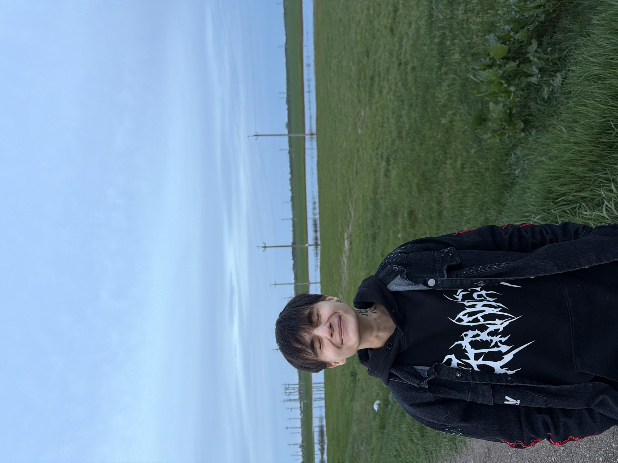 The image shows a person with short dark hair, smiling with eyes closed, wearing a black hoodie with a white graphic print and a black denim jacket with red details. The person has a tattoo on their neck.
The background consists of a sky with light blue hues, a green field, and electrical power lines. Azyakul', Russia, has experienced flooding; the water is visible between the green fields.