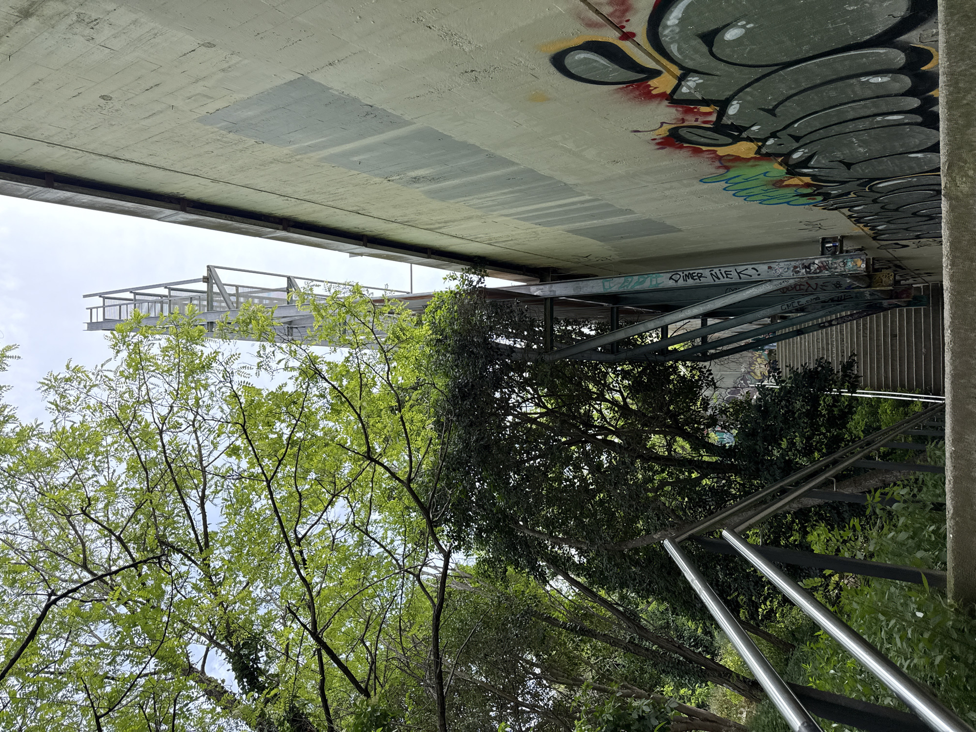 An upwards-angled shot presents a section of urban infrastructure interwoven with nature, taken in what appears to be the city of Barcelona, Spain. The perspective is dominated by the underside of a concrete bridge or overpass, which takes up the upper portion of the frame and bears graffiti. To the left, a metal walkway or viewing platform extends from the structure, supported by a framework of steel beams. A staircase ascends from the lower right, its silver metal railing clearly visible against the green foliage. Trees surround the staircase and walkway, their branches and leaves contributing a natural, organic element to the scene. The sky is visible through the gaps in the trees.