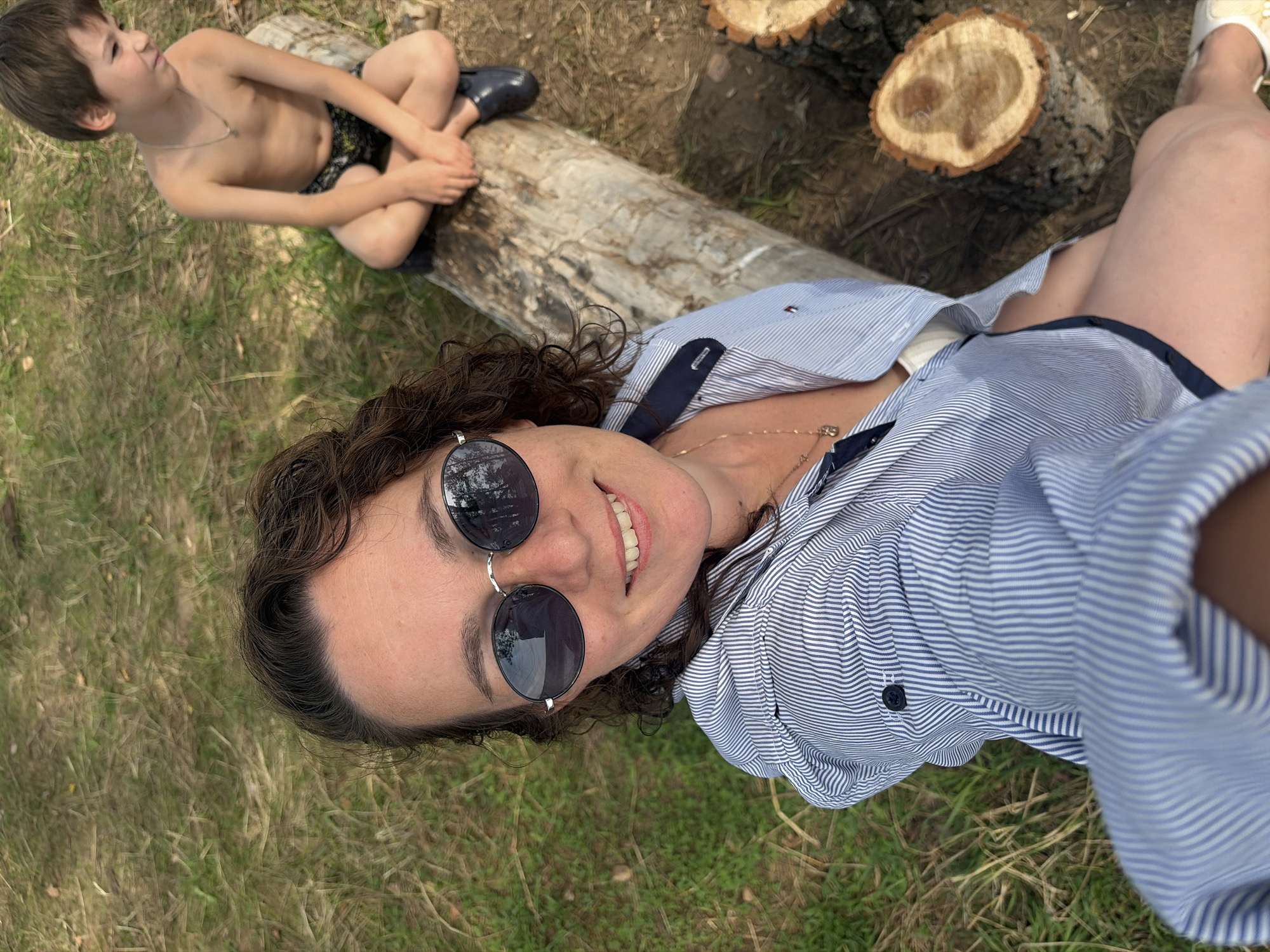A boy and a woman are outdoors, possibly in a park or a wooded area. The boy is shirtless, wearing dark shorts with a pattern, and has a silver chain around his neck. He is sitting on a large, fallen log, with his legs crossed. To the right of the boy, there are two cut tree stumps. The woman is lying down, in the foreground of the image. She is wearing a blue and white striped button-down shirt, with a dark collar and dark buttons. She is also wearing sunglasses with round, dark lenses and a thin silver frame. A thin gold necklace is visible around her neck. The ground around them is covered in dry grass and some patches of green grass. The overall lighting suggests it is daytime, with bright sunlight. The scene appears relaxed and casual, possibly a family outing. There is a small red tag with white lettering on the woman's shirt, near the collar, which is too blurry to read.