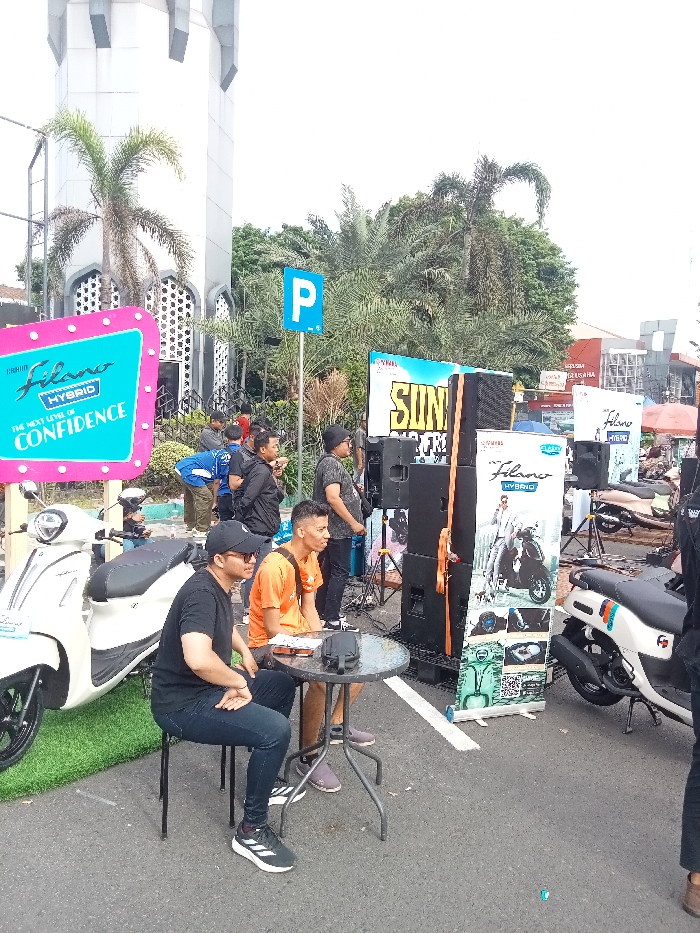 Outdoor scene shows a group of people near a parked scooter and promotional displays. Two men sit at a small round table. One wears a black shirt and baseball cap, the other an orange shirt. Behind them, others stand near audio speakers and a large sign for "Filano Hybrid". A white scooter is parked on the left. A blue parking sign is visible. Palm trees and a building with a white turret-like structure are in the background. The event seems to be a product promotion, likely for the scooter.