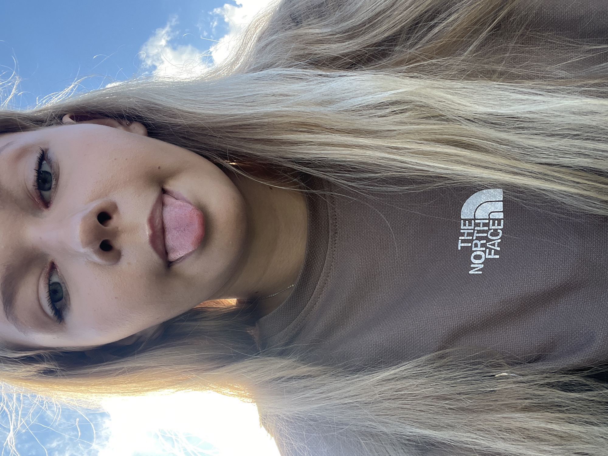 The image shows a young person with blonde hair outdoors against a bright blue sky with some clouds. The person is sticking their tongue out, and is wearing a brown sweatshirt with "THE NORTH FACE" logo. The lighting suggests it is daytime. The location is not specified in the image or in the provided context.