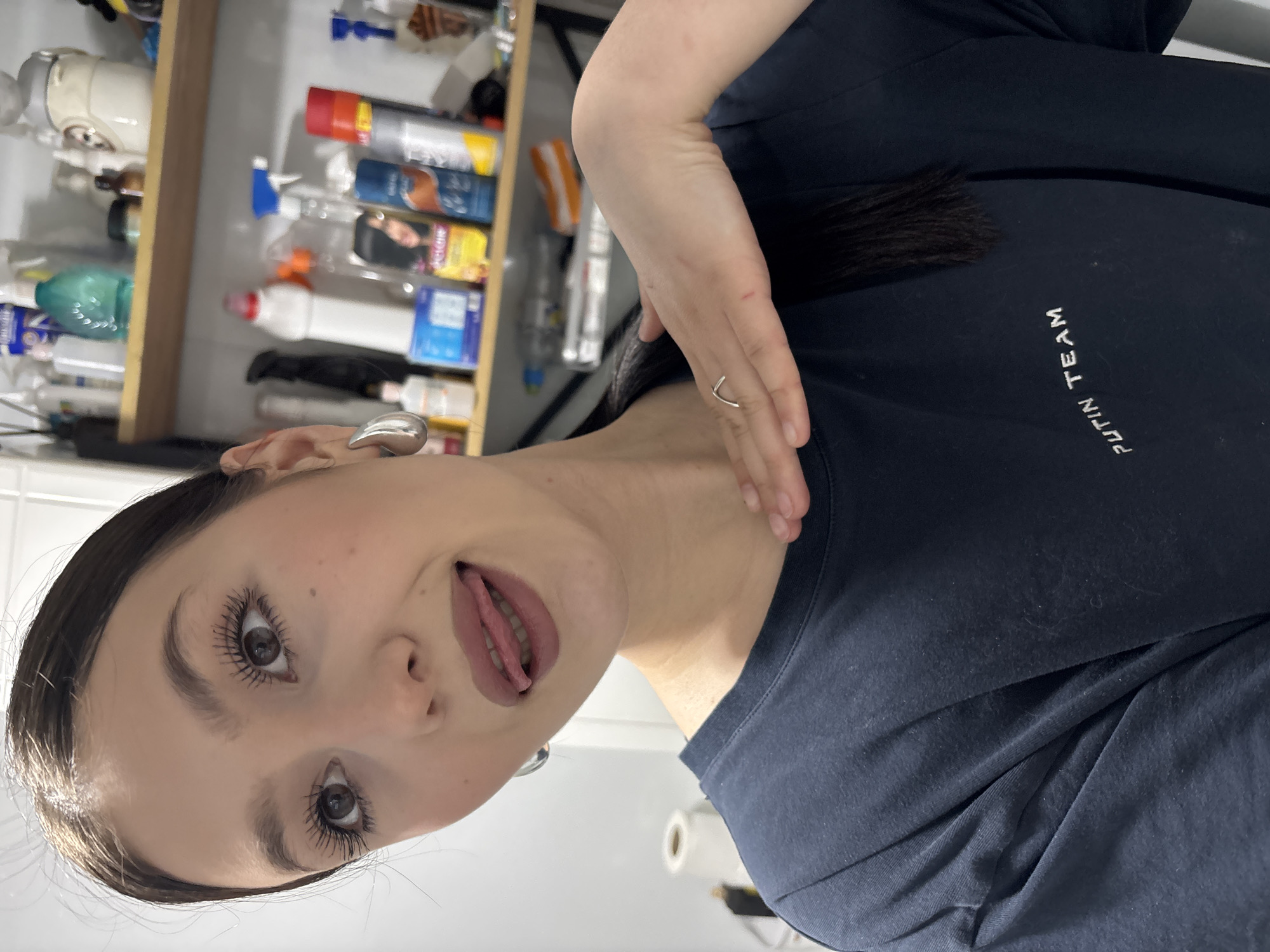 A woman with dark hair and fair skin is pictured in a close-up, tilted shot. Her tongue is slightly extended. She is wearing a dark blue t-shirt with white lettering that reads "PUTIN TEAM". The background includes a shelf filled with various household items and cleaning products, and a white tiled wall. The location cannot be determined, and there is no evidence of it being in the city of Фоки, Russia. There are no obvious actions or interactions taking place other than the woman posing for the photo.