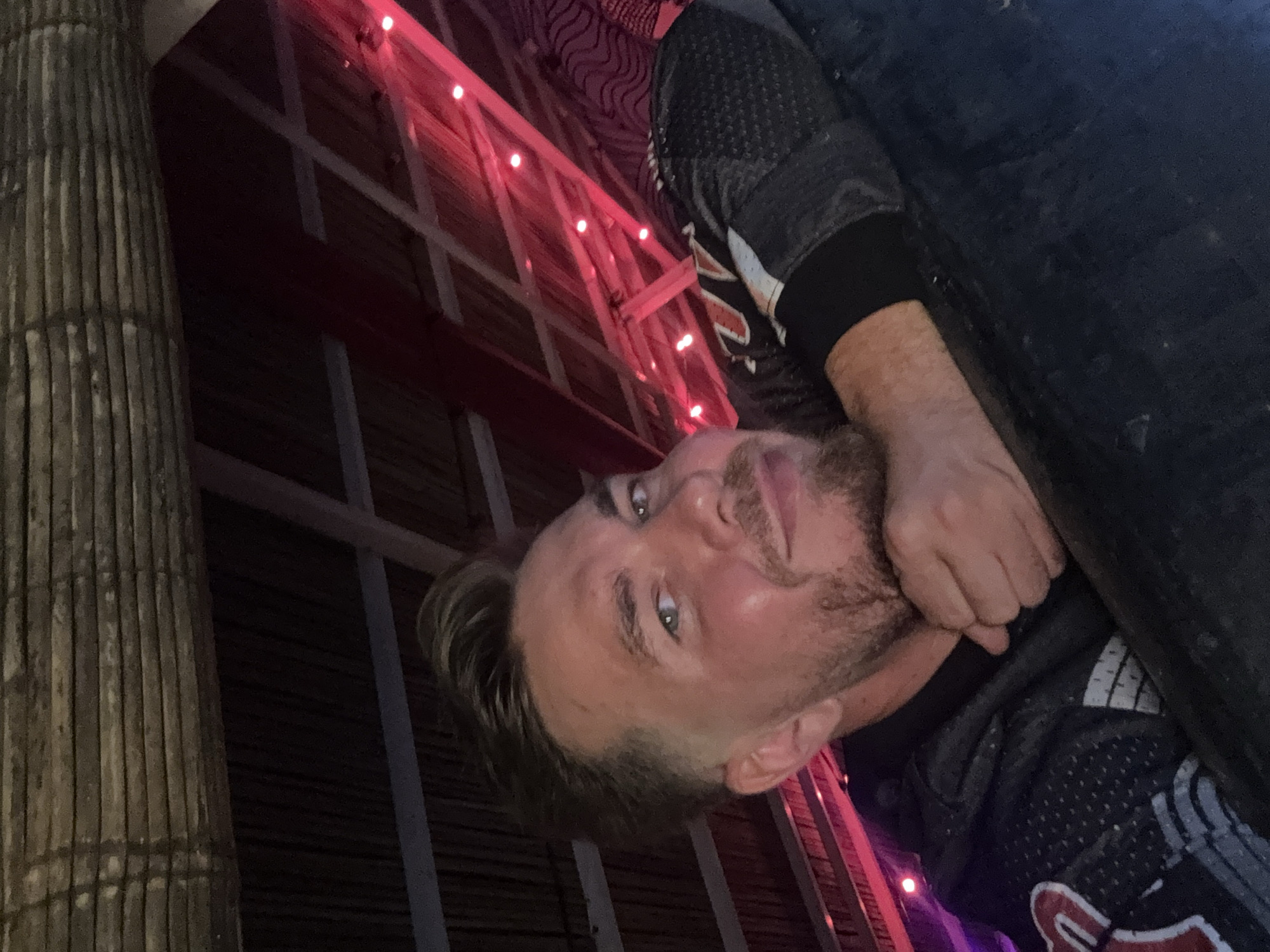 A Caucasian male with short blond hair, light stubble, and light eyes is posing for a photo. He's wearing a dark, patterned sports jersey with white and red accents. His right hand is near his neck. The background features a bamboo-like structure on the left, and a building with a red-lit framework. The image appears to be taken in Canggu, Indonesia.