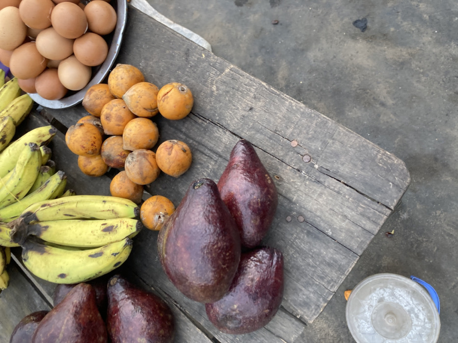 A wooden table displays various food items. A silver bowl contains brown eggs. Bunches of yellow bananas lie adjacent to small, round, orange fruits. Four dark, ripe avocados are arranged on the table's surface. A light gray bucket is placed near the table's edge. The backdrop consists of a light gray concrete surface.