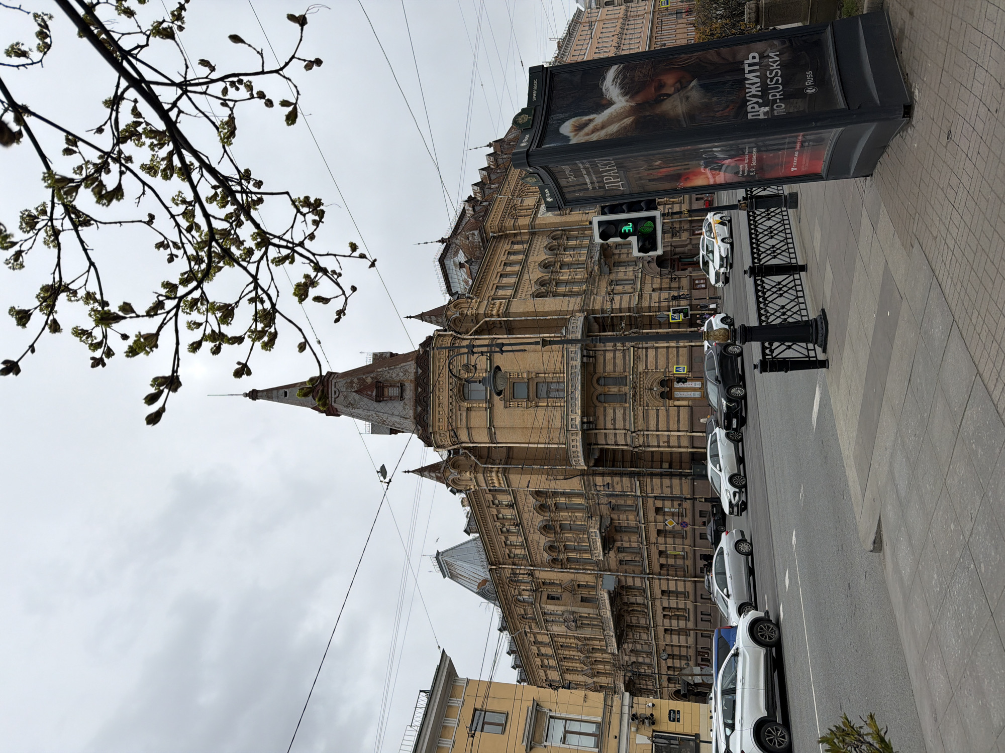 Here is a description of the image:
The image depicts a street scene in Saint Petersburg, Russia, featuring a distinctive architectural structure and urban elements. The building is ornate, with a complex facade including towers, windows, and decorative elements. There is a prominent sign on the street that advertises a show called "Dracula." In addition, one of the signs states "friend by Russki" and in other languages. A traffic light is on green. Several cars are parked along the side of the street next to a low, black fence. The sky is overcast. In the foreground, there are tree branches with buds. Power lines are visible crisscrossing the sky. The image is angled, with the top of the building pointed towards the left side of the image.