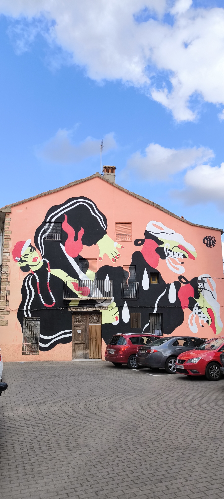 In Fanzara, Spain, a vibrant mural adorns the salmon-pink facade of a building. The artwork depicts stylized, abstract figures in black, pink, and yellow, characterized by bold outlines and simplified forms. One prominent figure on the left has green skin, red lips, and wears a beaded necklace, while another figure on the right has a skull-like face with "XXX" etched on its teeth. The mural spans the upper portion of the building, covering windows and a balcony. Below the mural, a weathered wooden door is centrally located, flanked by barred windows. The foreground features a paved courtyard with several cars parked, including a red hatchback, a silver sedan, and another red car. The sky above is a bright blue, dotted with fluffy white clouds.