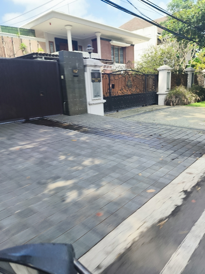 The image shows a paved driveway in front of a house located in Bandung, Indonesia, at the coordinates -6.879 latitude, 107.586 longitude. The driveway is made of gray rectangular tiles and is bordered by a white line. The house is a two-story building with a large front gate and a brown door. There is a white mailbox to the right of the door, and the gate is made of black metal with an ornate design. There is a small patch of green grass and bushes on the right side of the driveway. The driveway is shaded by the house, creating a pattern of light and shadow on the tiles. The street is visible in the foreground, and there is a vehicle's rearview mirror in the lower right corner. The image is taken from the point of view of a person looking down the driveway towards the house.