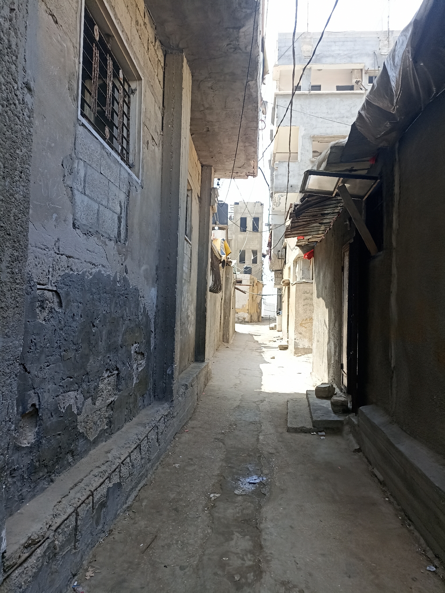 An alleyway in Gaza City, Palestinian Territories, is depicted in this image. The narrow passage is flanked by buildings constructed from concrete blocks and stucco, showing signs of wear and tear. On the left, a building features a window with bars and a rough stucco finish. To the right, another building has a rough concrete exterior and a sloped corrugated metal roof over a doorway. The ground of the alley is unpaved and uneven, with visible debris and a small puddle of water. In the distance, additional buildings are visible, some appearing unfinished or damaged, with electrical wires strung overhead. The scene is illuminated by sunlight, casting shadows that highlight the textures and imperfections of the environment.