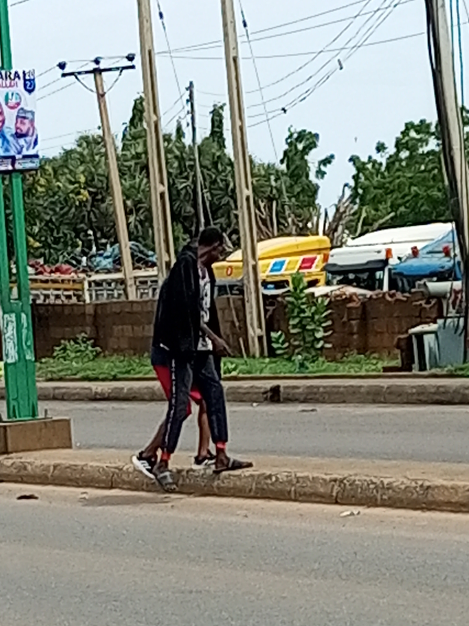 This image captures a street scene in Gombe, Nigeria, with two individuals walking on the sidewalk. The person in the foreground, facing away from the camera, is wearing a dark jacket over a white t-shirt with a graphic print and dark trousers with a stripe down the side. They are wearing black sandals with white accents. To their left and slightly behind, another individual is visible, wearing red shorts and dark trousers. The background features a row of trees, a brick wall, and several vehicles, including a yellow truck with blue and red markings and a white truck. Overhead, power lines stretch across the frame, supported by utility poles. A green pole with some white markings is present on the far left, and a political poster is affixed to it. The ground is paved with asphalt, and the sidewalk is bordered by a concrete curb.