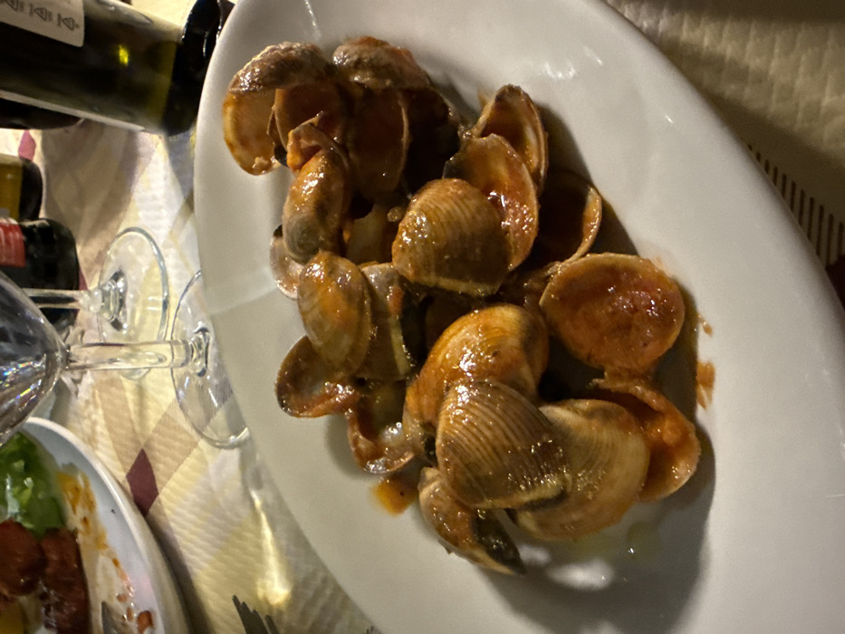 A close-up view depicts a white, oblong ceramic plate prominently featuring a serving of cooked clams (likely *vongole* or *cozze*) submerged in a reddish-orange, oil-based sauce. Approximately two dozen clam shells, many open and some closed, are arranged on the plate. To the upper left, a partial view of a second white ceramic plate reveals remnants of a prior course, including a brown, fried food item and green lettuce leaves. In the background, two clear stemmed wine glasses are visible, one positioned behind the other. The upper right corner shows the neck and shoulder of a dark green wine bottle, with another indistinct dark bottle behind it. The table is covered with a patterned tablecloth featuring light beige and maroon squares. Warm, artificial illumination is present, casting reflections on the glassware and the clam sauce. The specific location within Lecce, Italy, cannot be determined from the visual evidence.
