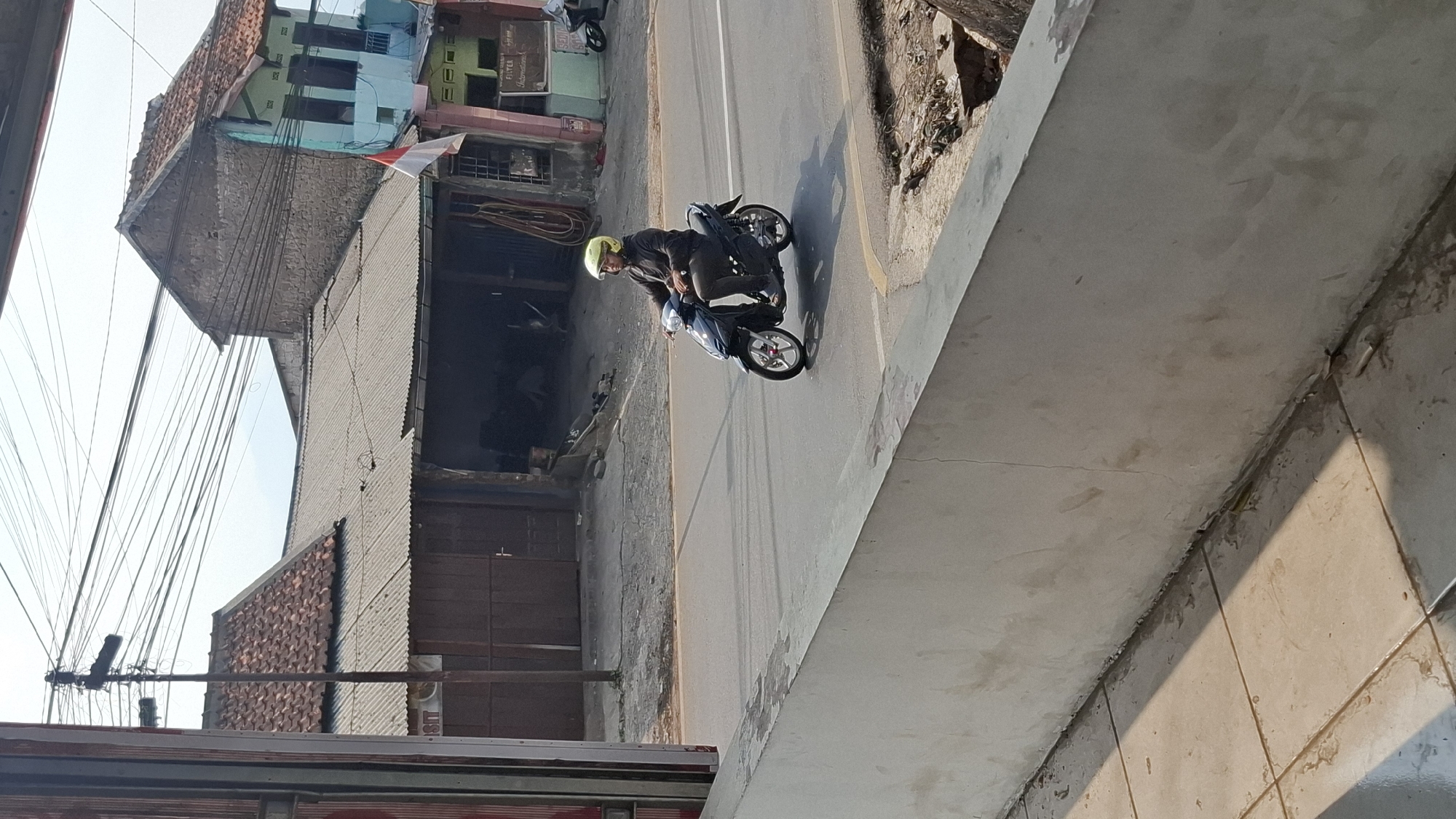 In the city of Cilaku, Indonesia, a person is riding a dark-colored scooter on a paved road. The rider is wearing a bright yellow helmet and a dark long-sleeved shirt with dark pants. The scooter has red accents on its wheels. The road is bordered by a light-colored concrete barrier in the foreground. Behind the road, there are several buildings with corrugated metal roofs and some with tiled roofs. One building on the right has a light blue facade and a red and white flag displayed. Power lines are visible crisscrossing the sky above the buildings. The overall scene depicts a street view in a residential or commercial area of the city on a bright, sunny day.