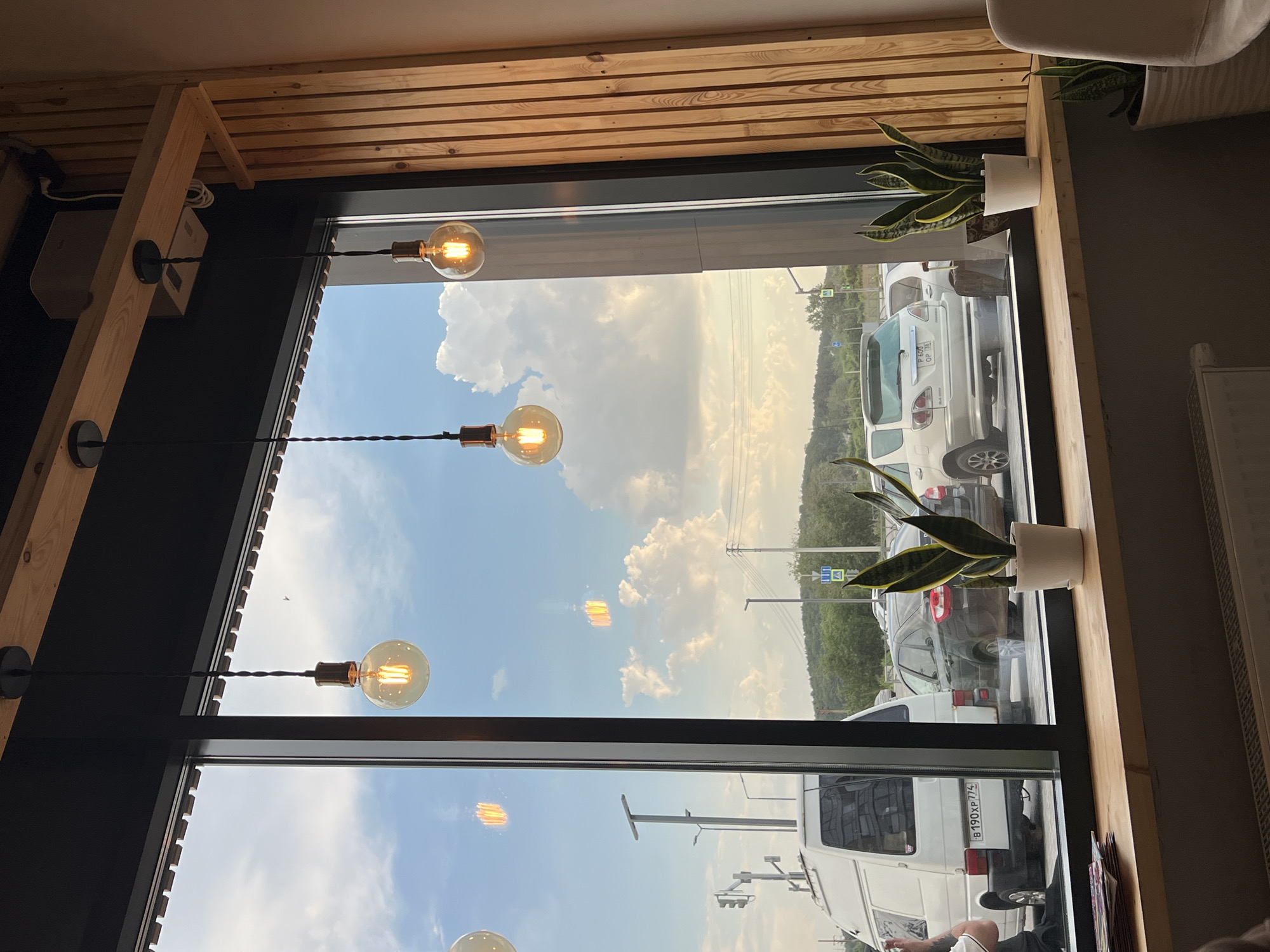 The image depicts an interior view through a large window, likely from a cafe or restaurant, looking out onto a street scene in Yekaterinburg, Russia. Three vintage-style Edison light bulbs hang from the ceiling, casting a warm glow. The window offers a view of parked cars, including a white van with a Russian license plate ending in "774" and a white SUV. Outside, a traffic light and street signs are visible, along with trees and a slightly overcast sky with cumulus clouds. On the interior windowsill, several snake plants in white pots are positioned. A stack of magazines rests on the left side of the windowsill. The overall aesthetic suggests a modern, cozy interior design with a focus on natural light and ambient lighting from the hanging bulbs.