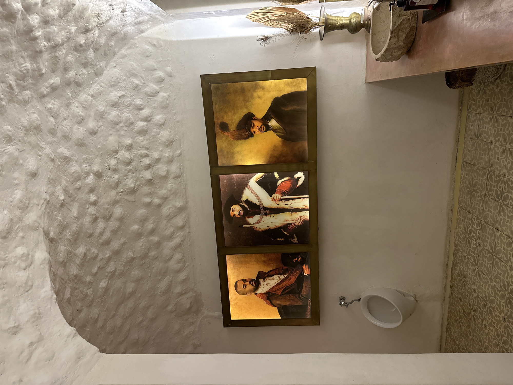 The image captures an eccentrically decorated restroom or washroom. On the left, a heavily textured, white stucco-like wall curves upwards, creating an organic, cave-like appearance.
The focal point is a vertical stack of three internally lit, framed portraits on the right wall. These artworks are styled as Old Master paintings but feature contemporary faces. The top portrait shows a man with a mustache and beard, wearing a dark cap with a feather, resembling Ice Cube, against a warm, golden background. The middle portrait depicts a man in a rich, fur-collared costume, possibly holding a staff or sword hilt. The bottom portrait features a bald man with a beard, wearing a red-brown jacket over a white shirt, resembling Kanye West, also against a golden background.
Below the portraits, a white ceramic urinal is mounted on the wall, accompanied by a silver-colored spigot. To the right of the urinal, the lower portion of the wall is covered in patterned beige and white tiles.
In the upper right corner, a rustic, reddish-brown shelf holds decorative items. These include a brass-colored urn or vase containing dried feathers and foliage, and a rough-hewn stone basin, likely a sink. The wall around this area is plain white.
The scene is static with no activity taking place. The indoor setting implies artificial lighting, with the illuminated portraits casting a warm glow. No visible text is present.
