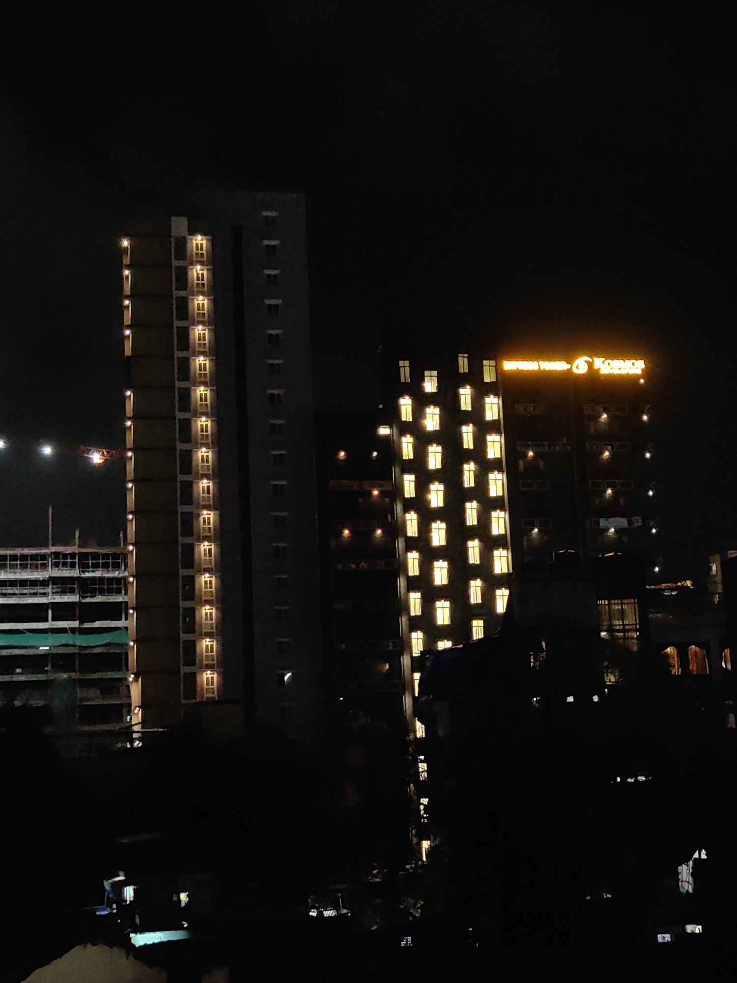 The image captures a nighttime view of a cityscape in Mumbai, India, featuring several high-rise buildings. In the foreground, silhouetted trees and structures are visible against the dark sky. To the left, a tall, narrow building with many illuminated windows stands out, its facade appearing to be a light brown color. Adjacent to it on the right is a wider, darker building with a similar pattern of illuminated windows. Further to the right, a taller, more prominent building displays a bright orange illuminated sign that reads "EMPRESS TOWERS" and "KOSMOS". This building also has numerous lit windows, some appearing to be square and others more elongated. To the far left of the frame, a construction site is partially visible with cranes and scaffolding against the night sky, suggesting ongoing development. The overall scene depicts a densely populated urban environment at night, characterized by illuminated architecture and the signs of continuous growth.