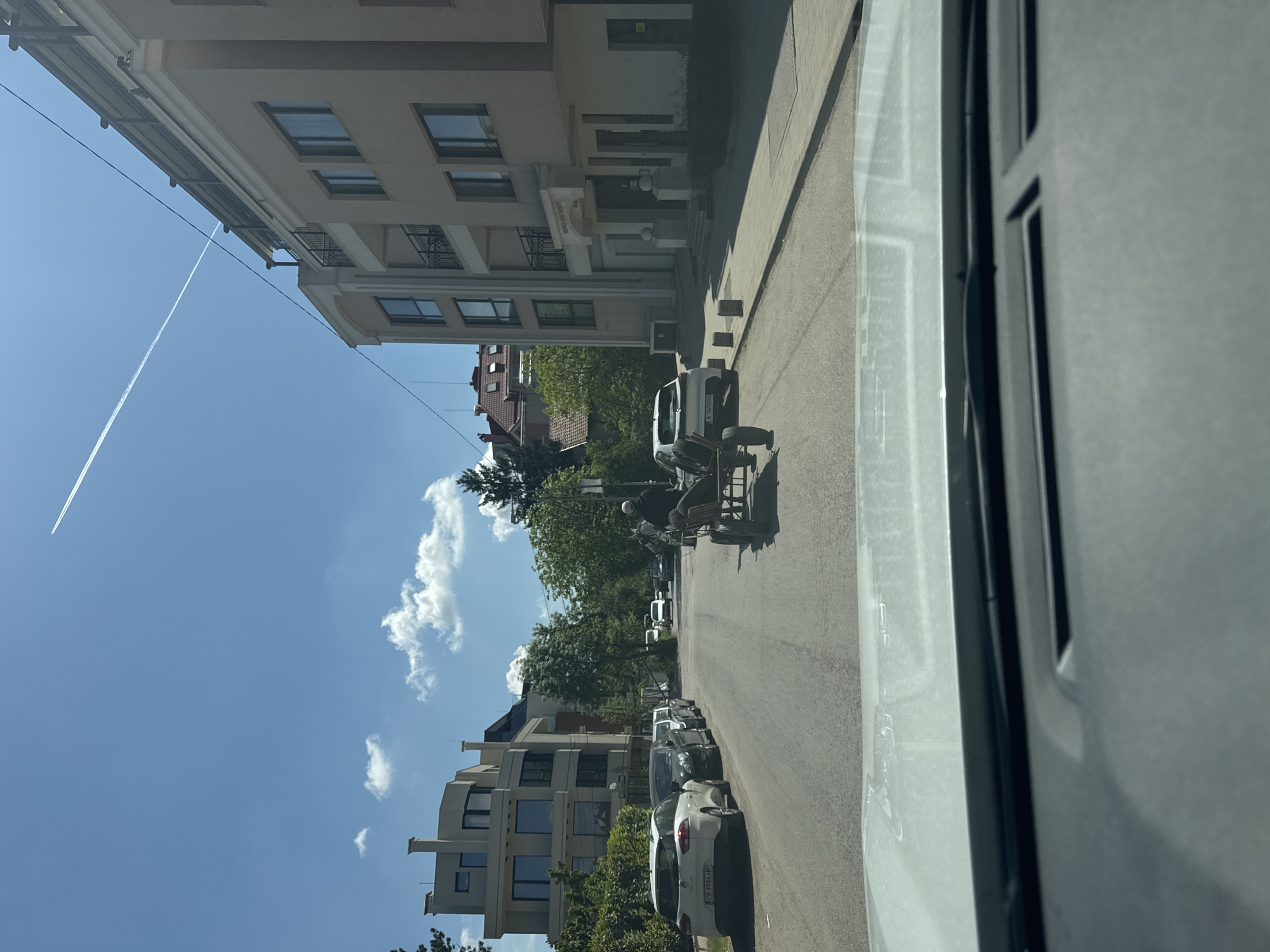 A slightly tilted medium shot taken from inside a vehicle, looking out onto a street lined with buildings and parked cars. Two men are working on what appears to be a utility cart in the middle of the street, closer to the foreground. The sky is blue with scattered clouds, and a contrail is visible. The buildings are multi-story, with residential-style architecture. Parked cars line the right side of the street. The scene is in Sofia, Bulgaria, based on the license plate of a white car parked on the street.