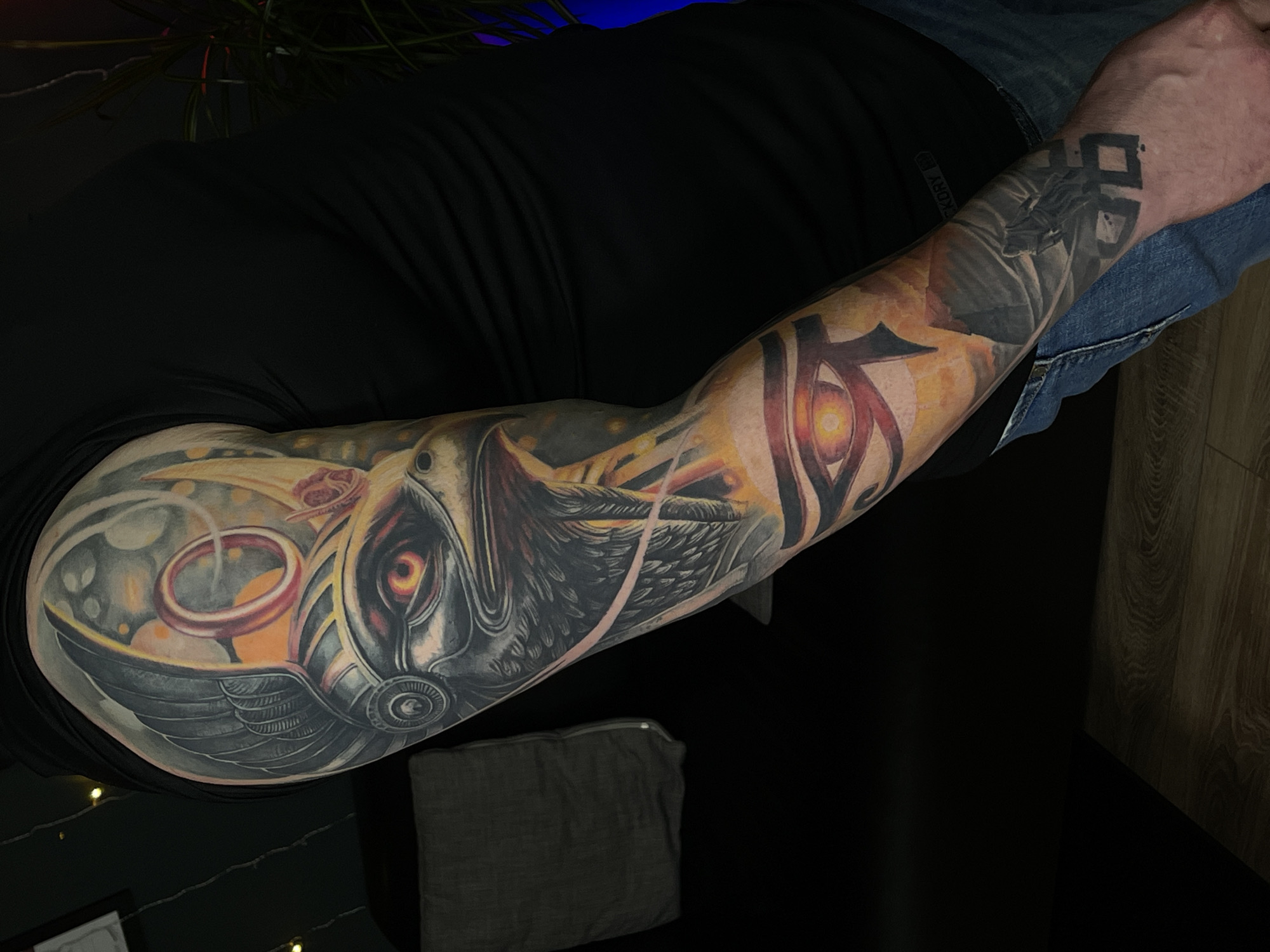 A man's arm is showcased in a photo, covered with elaborate tattoos. The arm features a black and grey tattoo of a mechanical bird or creature, with an eye that has an orange pupil. The design incorporates a golden ring element as well. Further up the arm, closer to the hand, there's an eye tattoo design using Egyptian symbols. At the wrist, there are angular black symbols tattooed. The man is wearing a black t-shirt and jeans. A grey zippered pouch is visible near his elbow. The background is dark, with some soft lighting and what looks like the corner of a wooden surface or floor. The location is not explicitly mentioned in the image details.