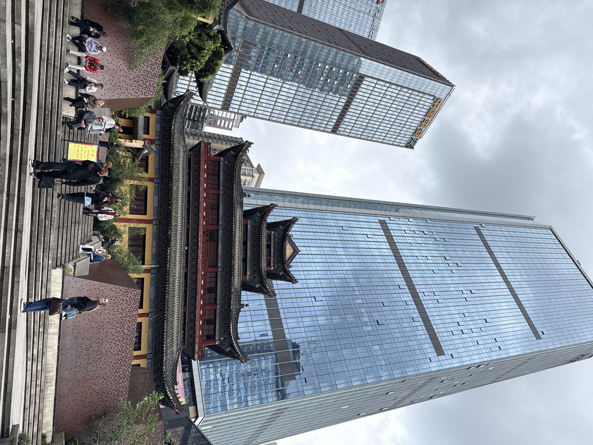 This urban scene in Yuzhong District, China, captures a compelling blend of traditional architecture and modern development under an overcast sky. The image, correctly oriented, shows a multi-tiered traditional Chinese building standing centrally, characterized by its ornate dark-tiled roofs, intricate carvings, and vibrant red structural elements.
To the left of the traditional building, a wide concrete staircase descends. Several people are casually seated on the steps, while others stand or walk. Near the bottom of the stairs, a person in black clothing is looking at a yellow rectangular sign displaying Chinese characters, including "渝中区" (Yuzhong District). Another person in jeans and a dark top walks down the stairs. Adjacent to the staircase and the traditional building is a modern structure featuring a reddish-brown facade with a distinctive geometric perforated pattern. Green foliage and trees are interspersed throughout this area, providing a natural contrast to the built environment.
In the background and extending to the right, two towering modern skyscrapers with reflective glass facades dominate the skyline. Their surfaces mirror the cloudy sky and fragments of neighboring buildings. On the upper section of the taller skyscraper on the right, golden Chinese characters are visible, spelling "中国农业银行" (Agricultural Bank of China). The scene illustrates the juxtaposition of historical cultural elements with contemporary urban expansion in a bustling city.