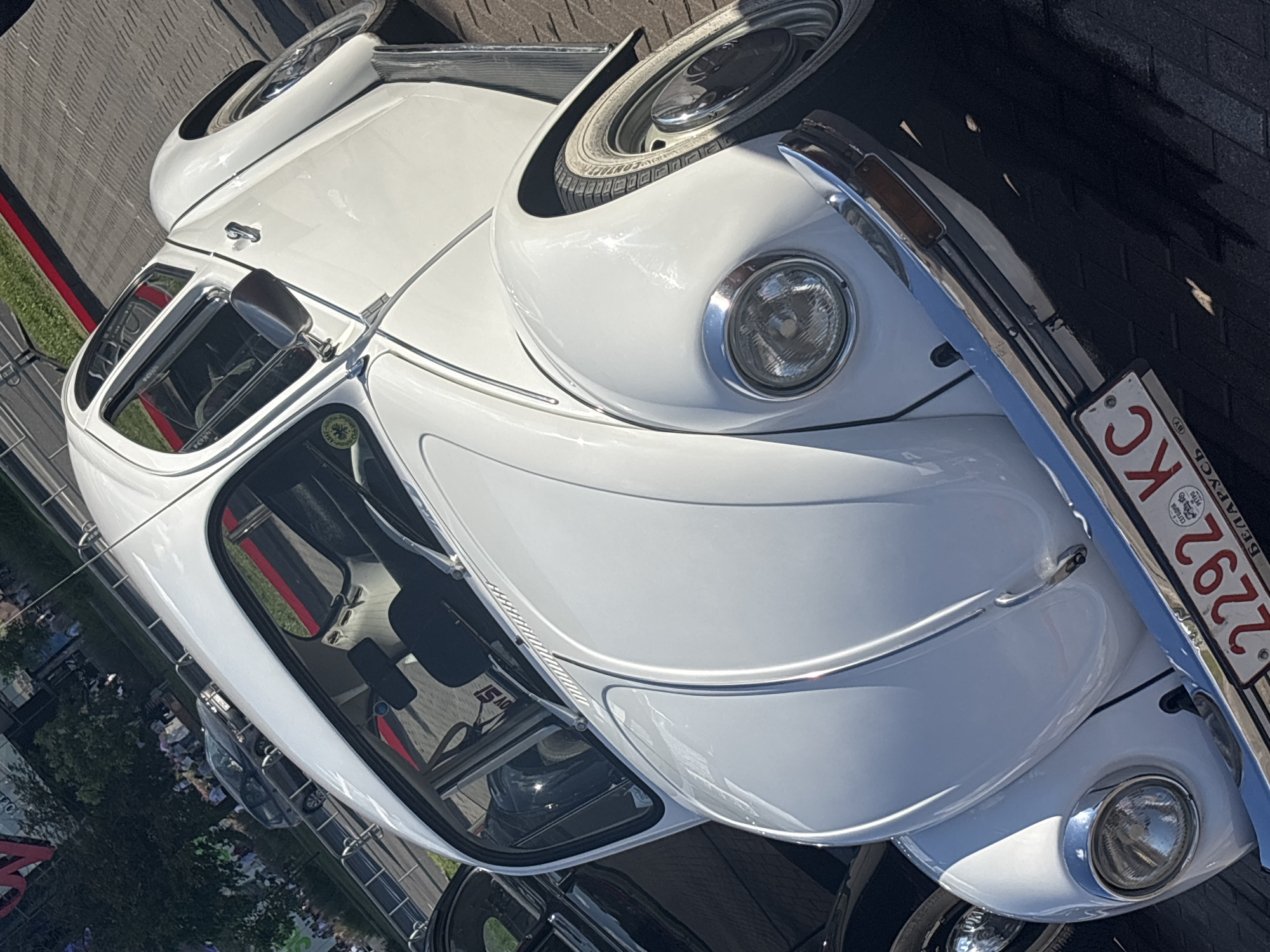 An eye-level, slightly tilted, close-up shot captures the front of a white Volkswagen Beetle on a paved surface. The car is the primary subject, with its chrome bumper, headlights, and front hood clearly visible. The license plate, which reads "2292 KC" and has "BY" on the bottom right corner, is mounted on the bumper. The car's white paint is reflective, showing some sunlight. To the left of the Beetle, a portion of a black car is visible. In the background, people and trees are present, suggesting an outdoor urban setting. The ground is paved with textured tiles, and a red line is visible on the pavement behind the car. The lighting suggests it is daytime, with clear visibility.