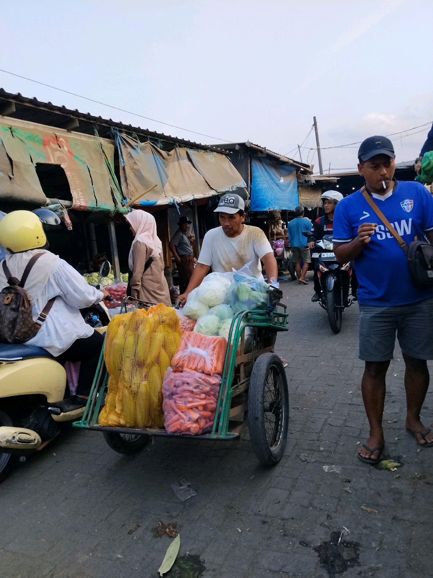 This is a candid, eye-level shot taken in a street market in Malang, Indonesia. The scene is bustling with activity and focused on a vegetable vendor pushing a cart laden with produce.
In the foreground, a man in a blue t-shirt and grey shorts, wearing a dark blue baseball cap, is prominently featured. He has a cigarette in his mouth and appears to be walking or standing near the cart. To his left, a vendor in a white t-shirt and a grey baseball cap is pushing a green cart filled with vegetables. The cart contains large sacks of corn and bags of carrots, along with some loose cabbage or other leafy greens.
To the left of the cart, a woman wearing a yellow helmet and a white long-sleeved shirt is seated on a yellow scooter, also carrying a backpack. Further in the background, a woman in a pink hijab and brown attire is seen conversing with someone partially obscured by the cart. Another man in a blue t-shirt stands further back, and a person on a motorcycle in a white helmet is also visible.
The market stalls are visible in the background, characterized by weathered tarpaulins and wooden structures. The ground is paved with cobblestones and shows signs of wear and tear, with some debris scattered around. The overall atmosphere suggests a typical, lively Indonesian street market.