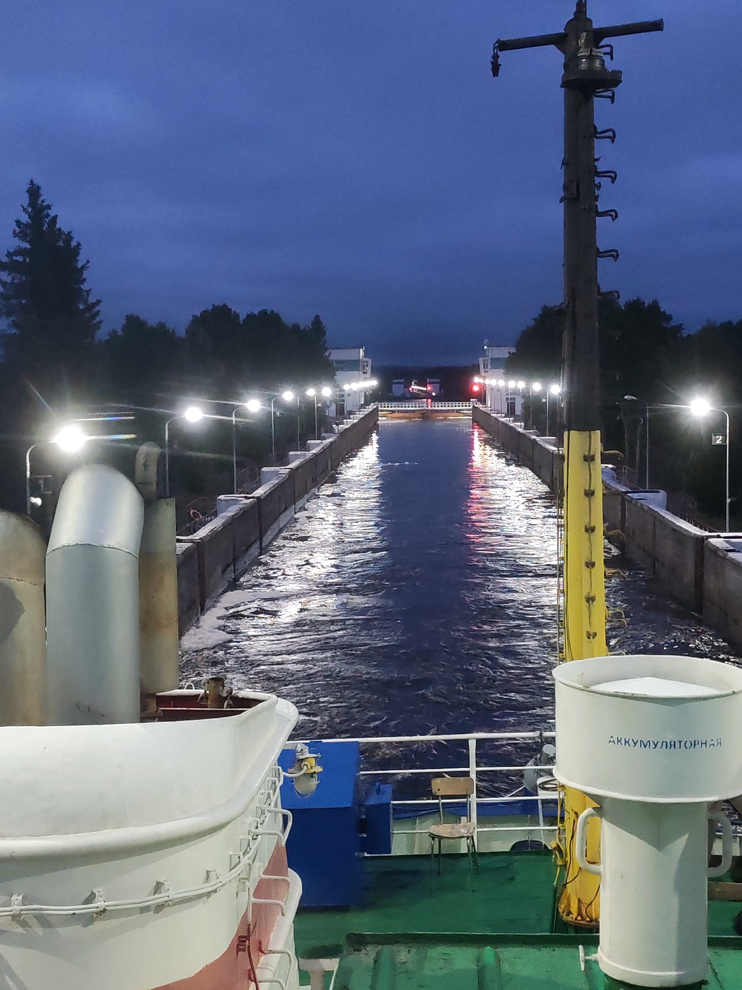The image shows a view from the deck of a ship as it enters a canal lock at dusk. The sky is a deep blue, transitioning to dark as night falls, and illuminated by a series of streetlights lining the canal walls. The canal is filled with turbulent water, indicating movement, and reflecting the lights. On the right side of the image, a tall, wooden pole with metal fixtures stands prominently, likely a guide or marker. In the foreground, the white and red hull of the ship is visible, along with a life raft and other deck equipment. The canal structure itself appears to be made of concrete, with metal railings and lights. Further down the canal, buildings and more lights are visible, suggesting an industrial or port area. The inscription on the white object in the foreground reads "АККУМУЛЯТОРНАЯ," which translates to "accumulator" or "battery room" in Russian. The location is specified as the city of Markovo, Russia. There are no people clearly visible in the image.