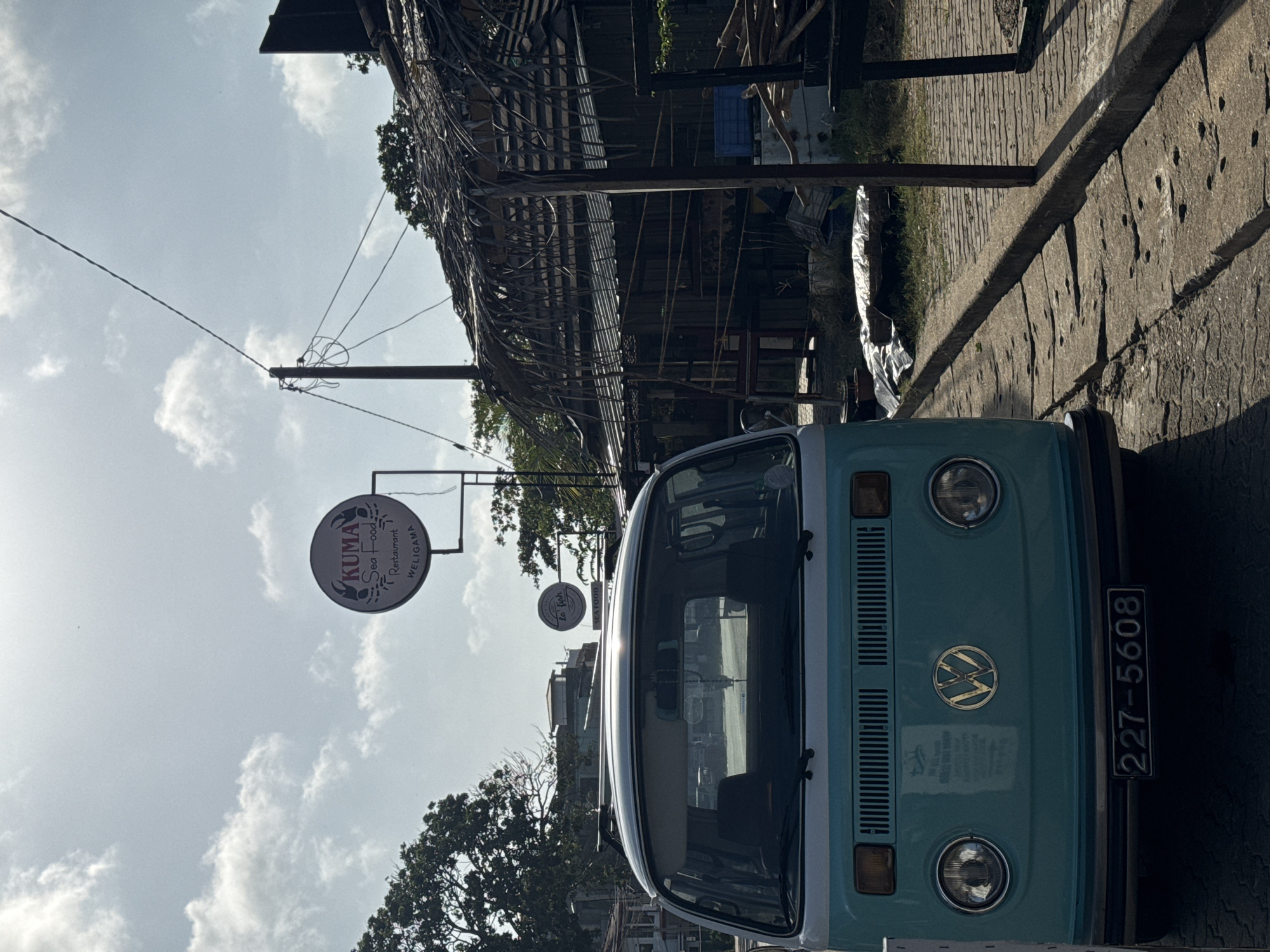 A front view of a teal-colored Volkswagen van is parked on a stone-paved street in Weligama, Sri Lanka. The van's license plate reads "227-5608." Behind the van, a sign for "KUMA Sea Food Restaurant" is visible against a bright, cloudy sky. Further in the background, a building with a thatched roof and wooden structures is present. Trees and utility poles are also part of the environment. The scene appears to be outdoors during daylight hours.