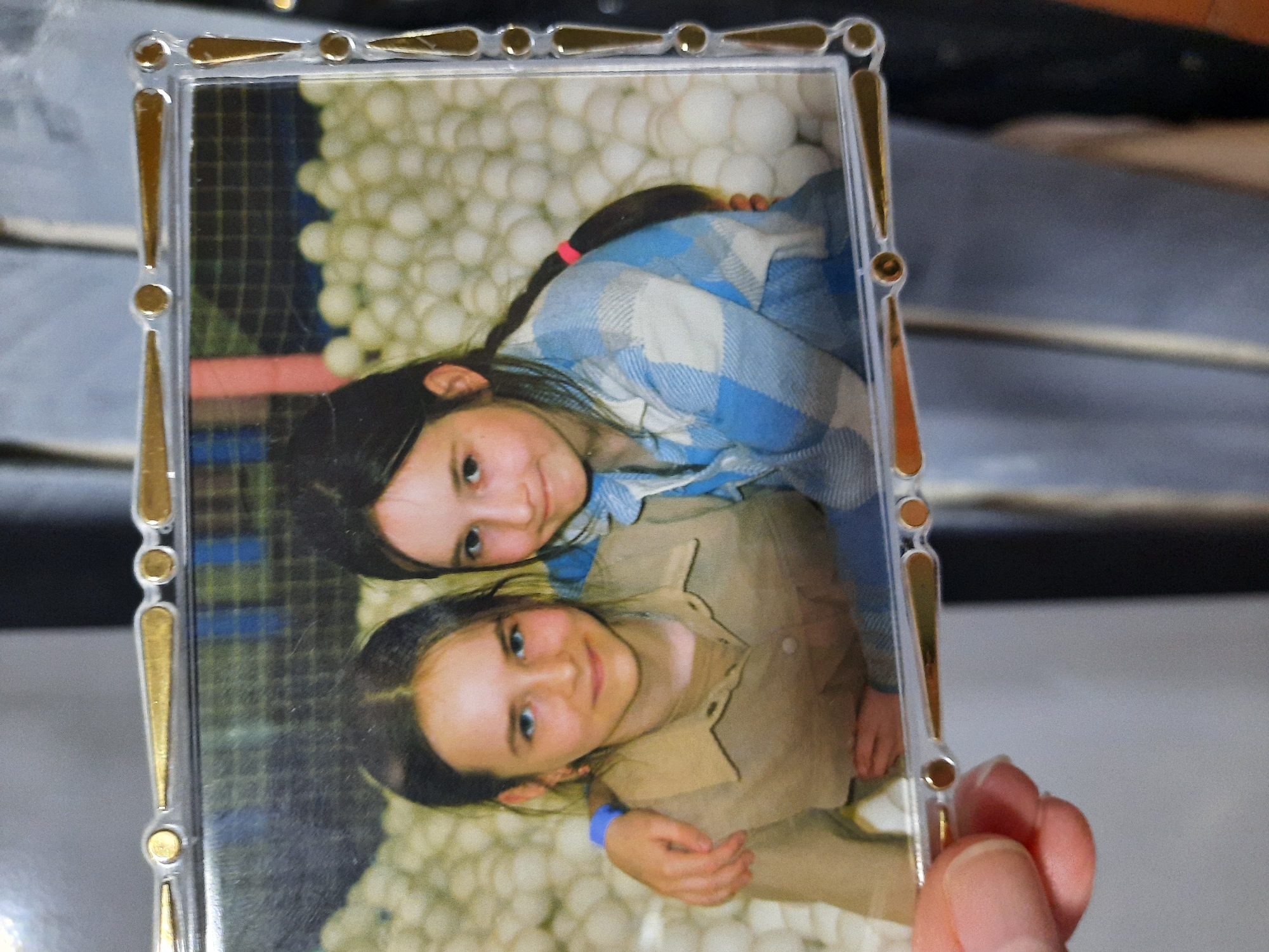The image is a photograph held in a clear plastic frame with gold accents. Inside the frame is a picture of two young girls embracing, seemingly in a ball pit filled with white balls.
The girl on top has dark hair pulled back in a braid with a pink hair tie. She's wearing a blue and white plaid shirt. The girl on the bottom has dark hair and is wearing a tan button-down shirt with a blue bracelet on her wrist. They are both looking at the camera and smiling.
The photo's background is entirely filled with white balls. Part of a human hand can be seen holding the frame. The background behind the frame is blurry and difficult to discern. There is no indication of Krasnogorskoye, Russia.