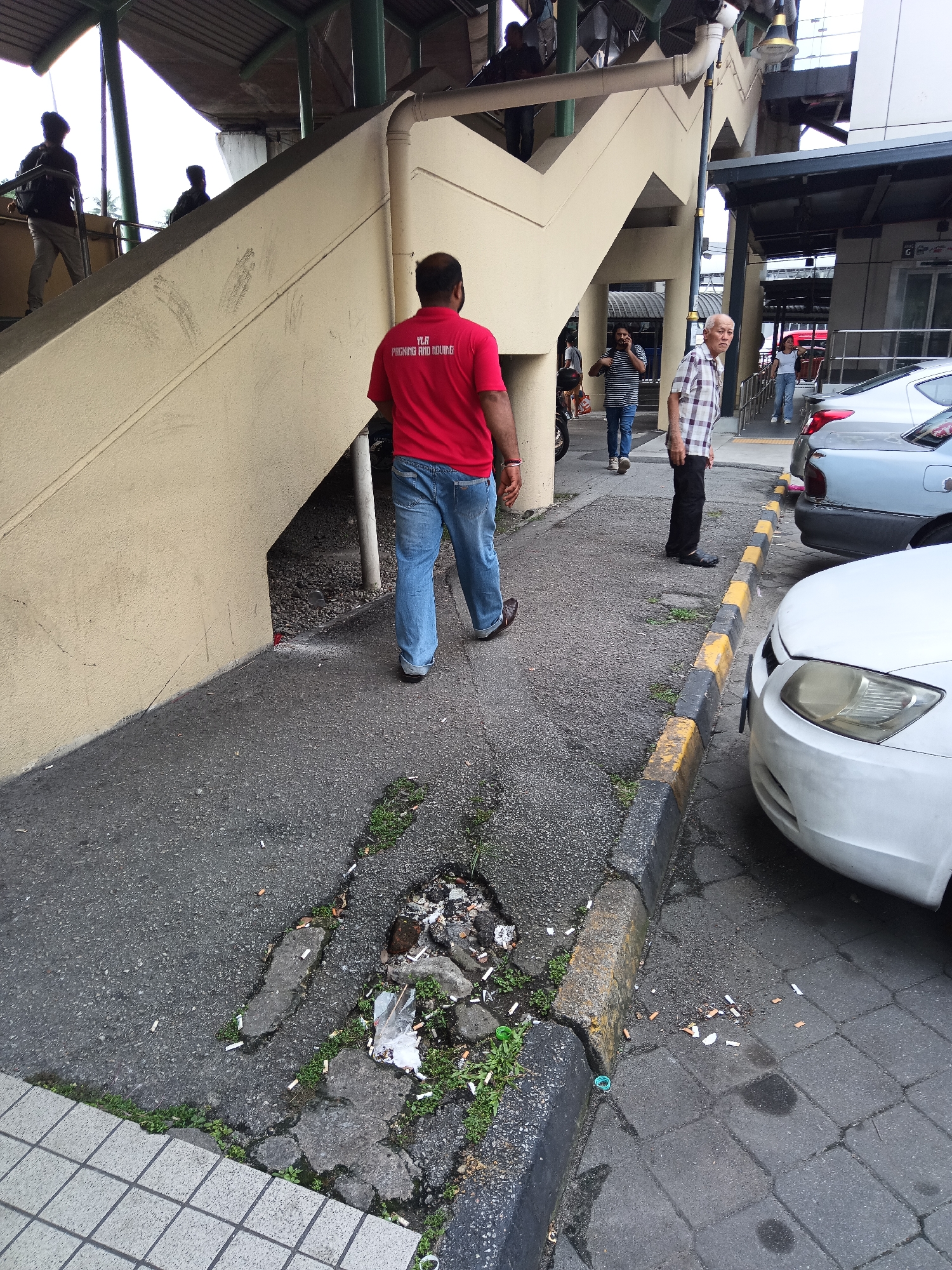 An overhead shot shows a man in a red shirt and blue jeans walking away from the camera down a cracked asphalt pathway in Kuala Lumpur, Malaysia. The man's shirt has text that reads "YLA PENGURUSAN BANGUNAN". He is walking towards a pile of garbage and cigarette butts in a large pothole on the pathway. To the right of the pathway is a yellow and black striped concrete curb, with a white car parked next to it. In the background, there are several people walking up a cream-colored concrete staircase that leads to an elevated walkway. The walkway is covered by a metal roof supported by green poles. The area appears to be an outdoor public space, possibly near a transportation hub, given the elevated walkway and the presence of parked cars.