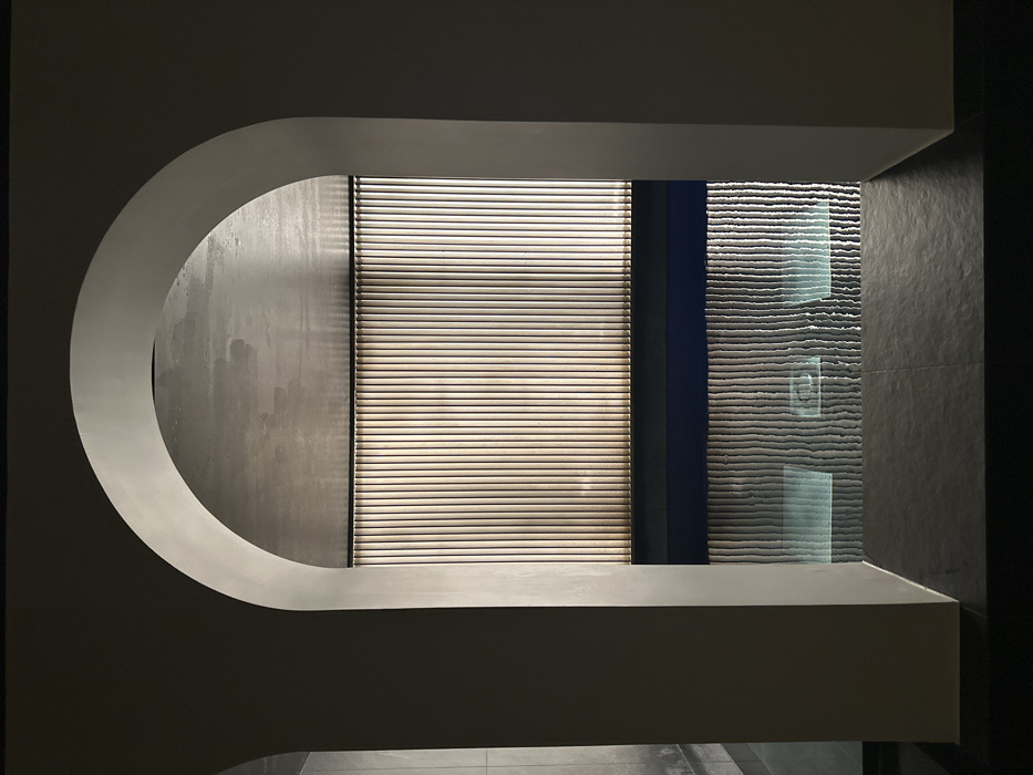 The image displays an interior view of the swimming pool area at the Hôtel Bvlgari Paris, France. A large, light-grey plaster archway frames the central scene, with its inner surface in shadow. Beyond the arch, a dark-tiled floor leads to a rectangular indoor swimming pool filled with dark blue water. The far wall of the pool area features a sequence of vertical, light-colored slats or panels, which are brightly illuminated, creating a strong vertical pattern of light and shadow. The surface of the pool reflects this slatted wall, with ripples causing distortion in the reflection. Multiple square, glowing light fixtures are visible submerged on the pool floor, their luminescence also reflected on the water's surface. The ceiling above the pool area appears to be a textured, light grey material. The scene is devoid of individuals, actions, or interactions.