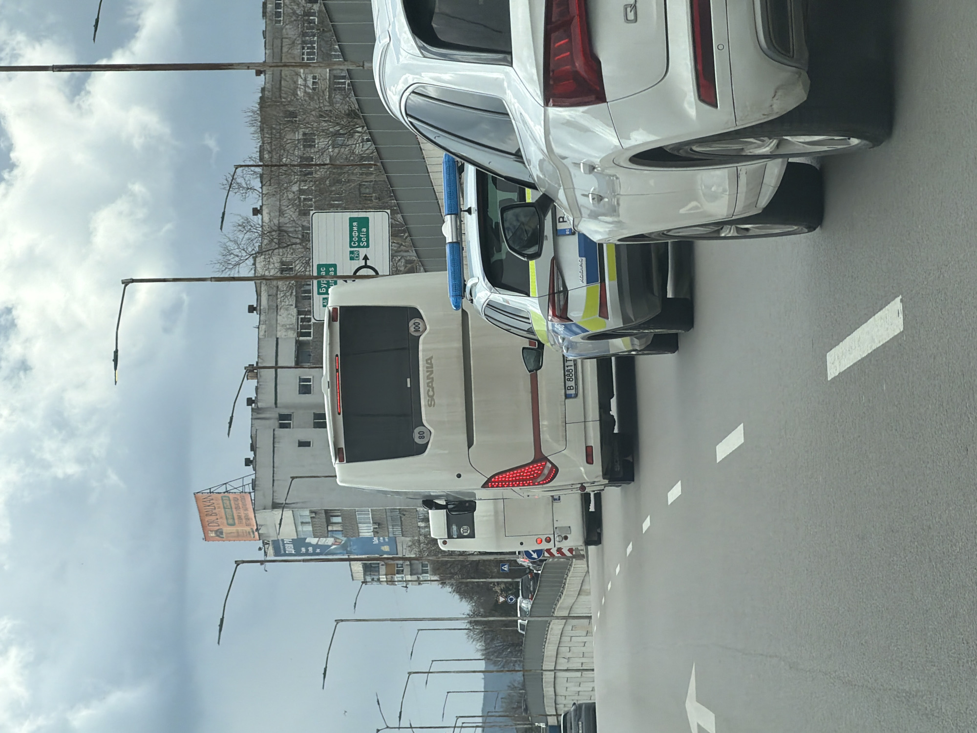 The image depicts a street scene, possibly taken from a vehicle. The focus is on the rear of several vehicles: a white SUV in the foreground, followed by a Bulgarian police car ("KODIAQ"), and a white Scania bus with "80" and "100" displayed on the back. Street signs indicate directions to Burgas and Sofia. The environment consists of urban buildings, streetlights, overhead signs, and cloudy skies. A billboard advertising "DR. BALKAN" is visible. The vehicles are positioned on an asphalt road marked with white lane indicators. Given the highway signs directing to Burgas and Sofia (cities in Bulgaria) and the license plate of the police car indicating Bulgaria, this scene is located in Bulgaria, specifically in the city of Varna.
