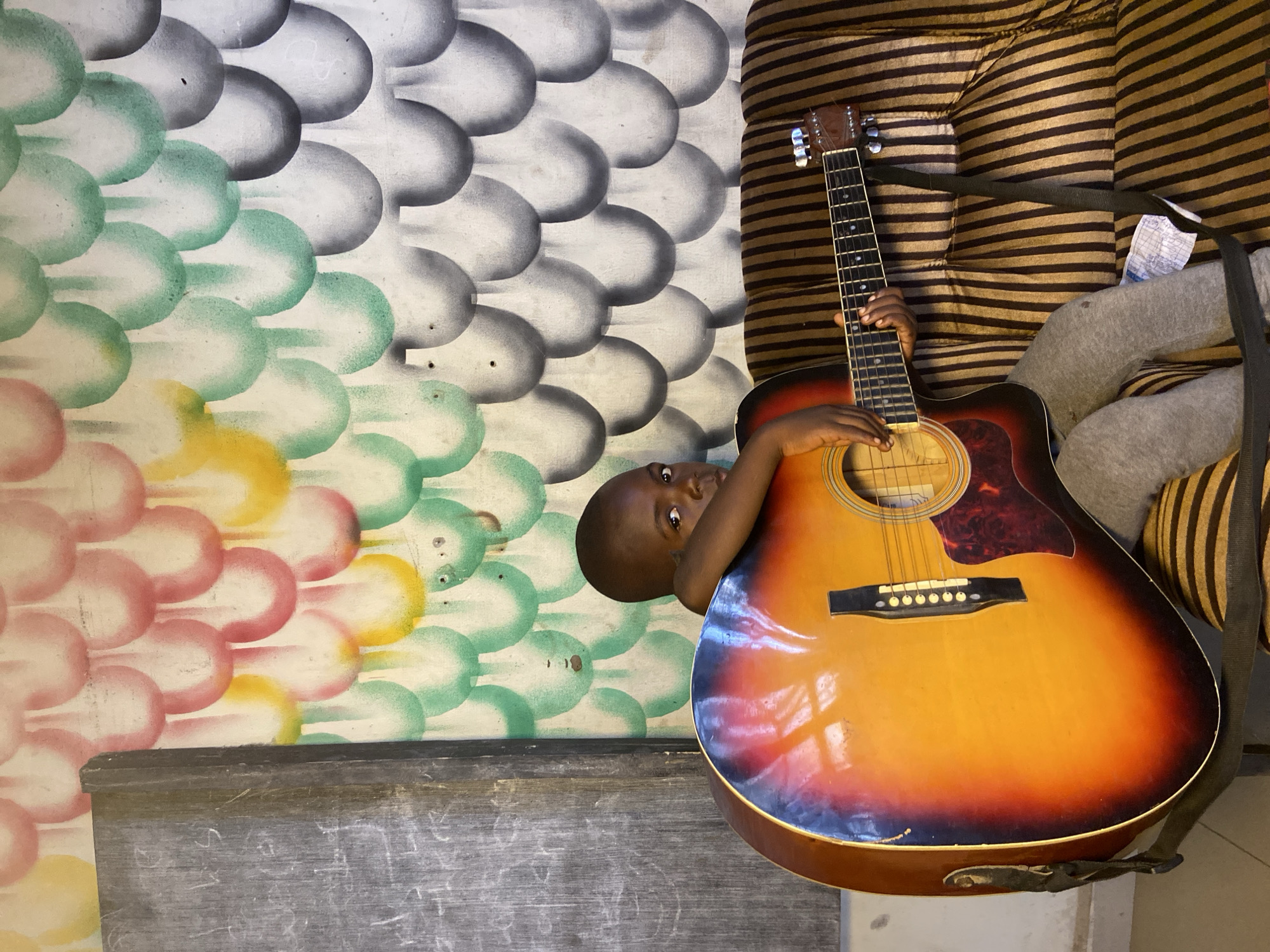 A child is visible with a partial view of their face, appearing next to an acoustic guitar with a sunburst finish. The guitar's headstock points upward. The child's hand is on the guitar's neck. A striped cushion is adjacent to the guitar. The background features a wall mural with rows of colored circles in shades of grey, green, yellow, and pink. A grey surface, likely a table or chalkboard, is at the bottom of the frame. The location is Mararaba, Nigeria.