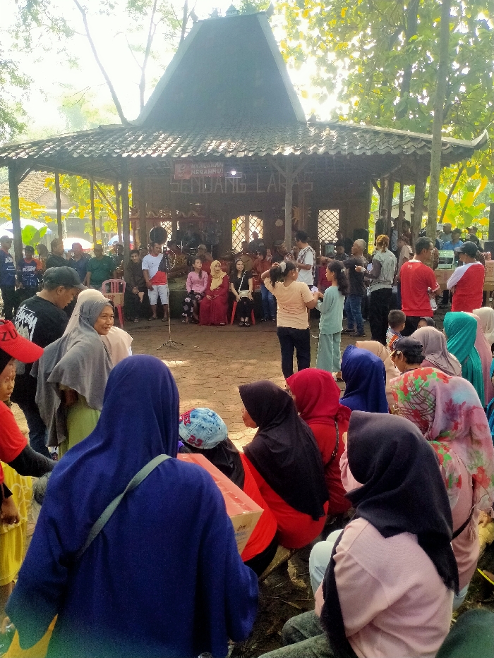 The scene is a gathering of people under a roofed structure, possibly a community hall, located in Masaran, Indonesia at coordinates -7.472 latitude, 110.956 longitude. The structure is made of wood and has a tiled roof. There are wooden pillars supporting the roof. In the foreground, a group of people are sitting on the ground, many wearing traditional clothing, including headscarves and hijabs. A few are wearing modern clothing. One individual is wearing a blue jacket with a black strap. The background shows a group of people standing, some of whom are interacting with each other. The ground is covered with reddish-brown soil. There are trees surrounding the structure. The air is bright and the scene is illuminated by natural sunlight. There is a sign on the wall of the structure with writing on it.