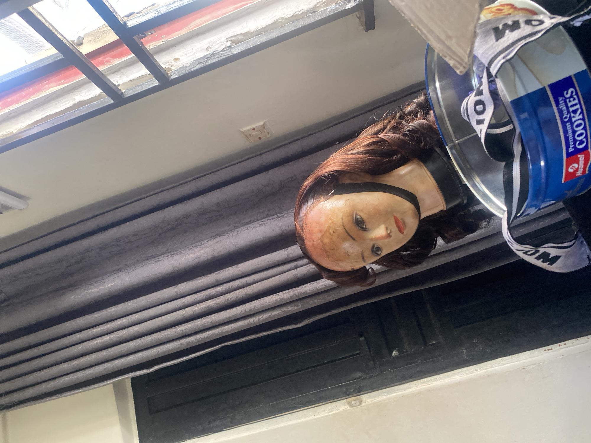 A mannequin head wearing a wig sits in front of a dark grey curtain and a dark wooden door in what appears to be a room in Kubwa, Nigeria. To the right of the mannequin, a window with a black metal grate reveals a glimpse of the outdoors, with some peeling paint on the window frame. Below the mannequin's head, a blue metal tin labeled "Premium Quality Cookies" is visible, with a black and white striped ribbon peeking out from inside it. The mannequin has light brown hair styled in waves, and is positioned at an angle to the camera. The lighting in the room suggests it is daytime.