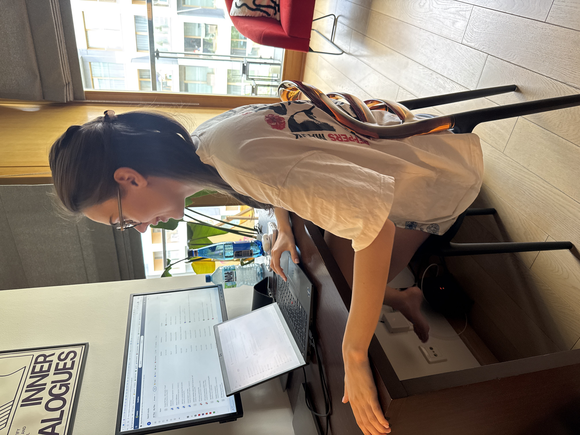 A woman is sitting at a desk working on a laptop with two computer monitors. She has brown hair pulled back and is wearing glasses and a white t-shirt. The desk is dark brown and cluttered with a keyboard, water bottles, and various objects. A black bag or container is visible under the desk. In the background, there is a red chair and a window showing a view of buildings across the street. A framed artwork hangs on the wall to the left of the monitors. The floor is light-colored hardwood. The location is Unknown, Unknown.