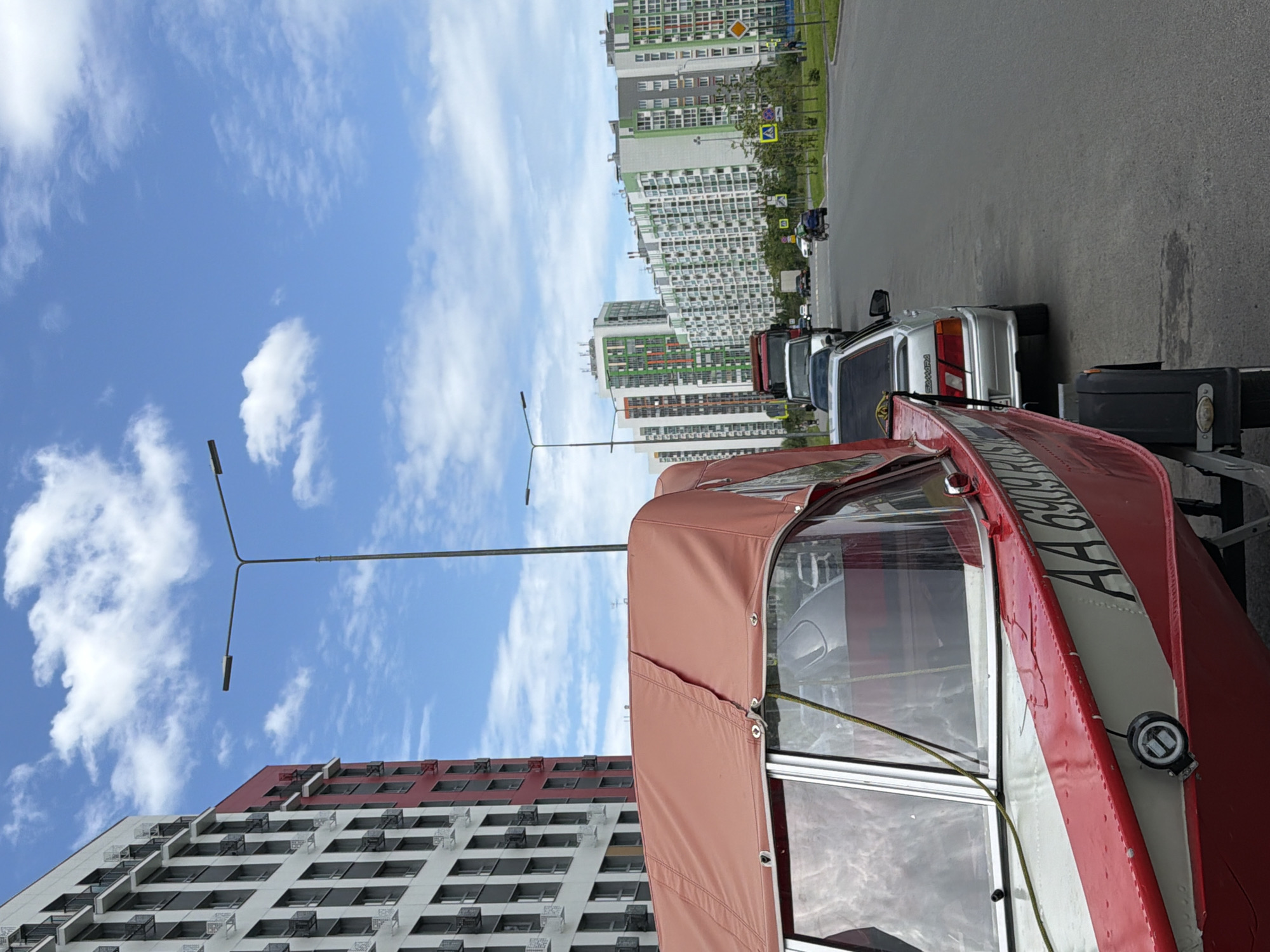 An eye-level, medium shot captures a red and white boat on a trailer parked on the side of a road. The boat has a tan canopy and clear windows, with the registration number "AA 6909 RUS" visible on its hull. To the right of the boat, several cars are parked in a line on the asphalt, including a silver sedan and a red truck. In the background, a row of multi-story apartment buildings with colorful facades stretches across the landscape under a partly cloudy blue sky. A street lamp stands on the left side of the frame. The environment appears to be a residential area in a city.