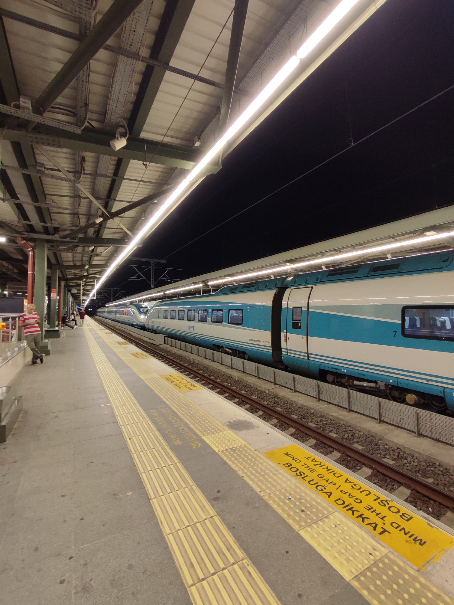 The image depicts a nighttime scene at a train station platform in Kadıköy, Turkey. A long, modern train with turquoise and white livery is stationary at the platform. The train's carriages are illuminated from within, and exterior lighting on the platform casts a glow on the scene. The platform itself is paved with concrete, featuring tactile paving strips and yellow warning signs in Turkish and English that read "MIND THE GAP" and "BOŞLUĞA DİKKAT." A few individuals are visible on the platform; one man in a striped shirt and trousers stands near the edge, leaning against a support pillar, while other passengers are seated or standing further down the platform. The station roof, supported by a metal structure, is visible overhead, with fluorescent lights illuminating the area. The tracks run alongside the platform, with gravel ballast visible. The background beyond the train is dark, indicating it is nighttime.