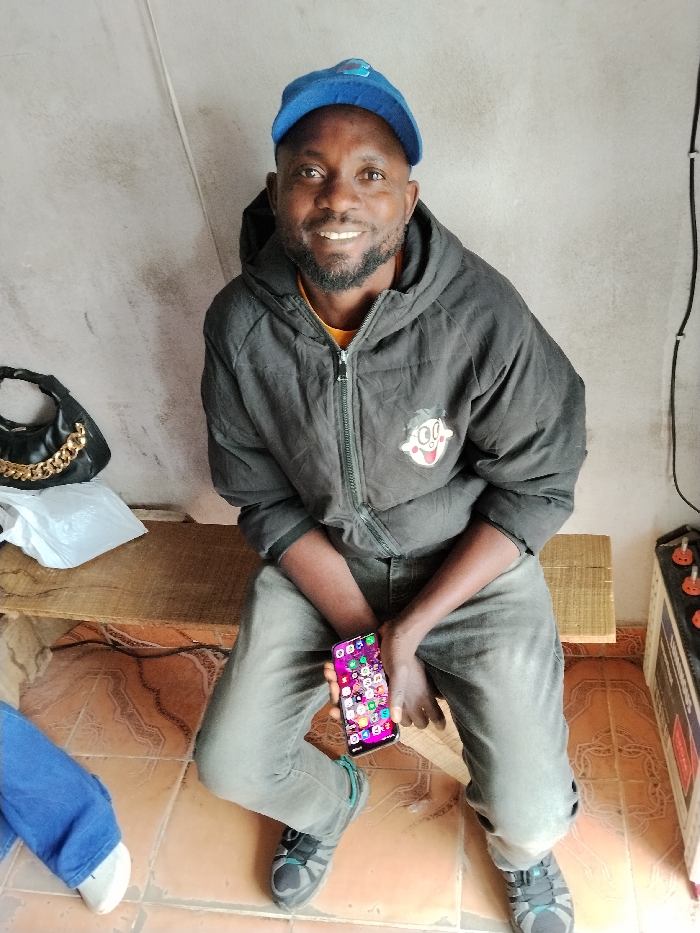 An adult male with dark skin and a beard smiles directly at the camera while seated indoors in Fagge A, Nigeria. He wears a blue baseball cap, a dark grey hooded jacket with a cartoon character emblem on the right chest, a yellow undershirt, and grey trousers. In his left hand, he holds a dark smartphone displaying a colorful grid of application icons. He is positioned on a light brown wooden bench. To his left on the bench, a black handbag with a gold chain strap and a white plastic bag are visible. The immediate background consists of a light grey wall and a light brown tiled floor with a repeating geometric pattern. On the far right, a partial view of a power unit featuring orange-capped terminals and black electrical wiring running vertically on the wall is observable. A lower leg in blue trousers and a white shoe is partially visible on the far left.