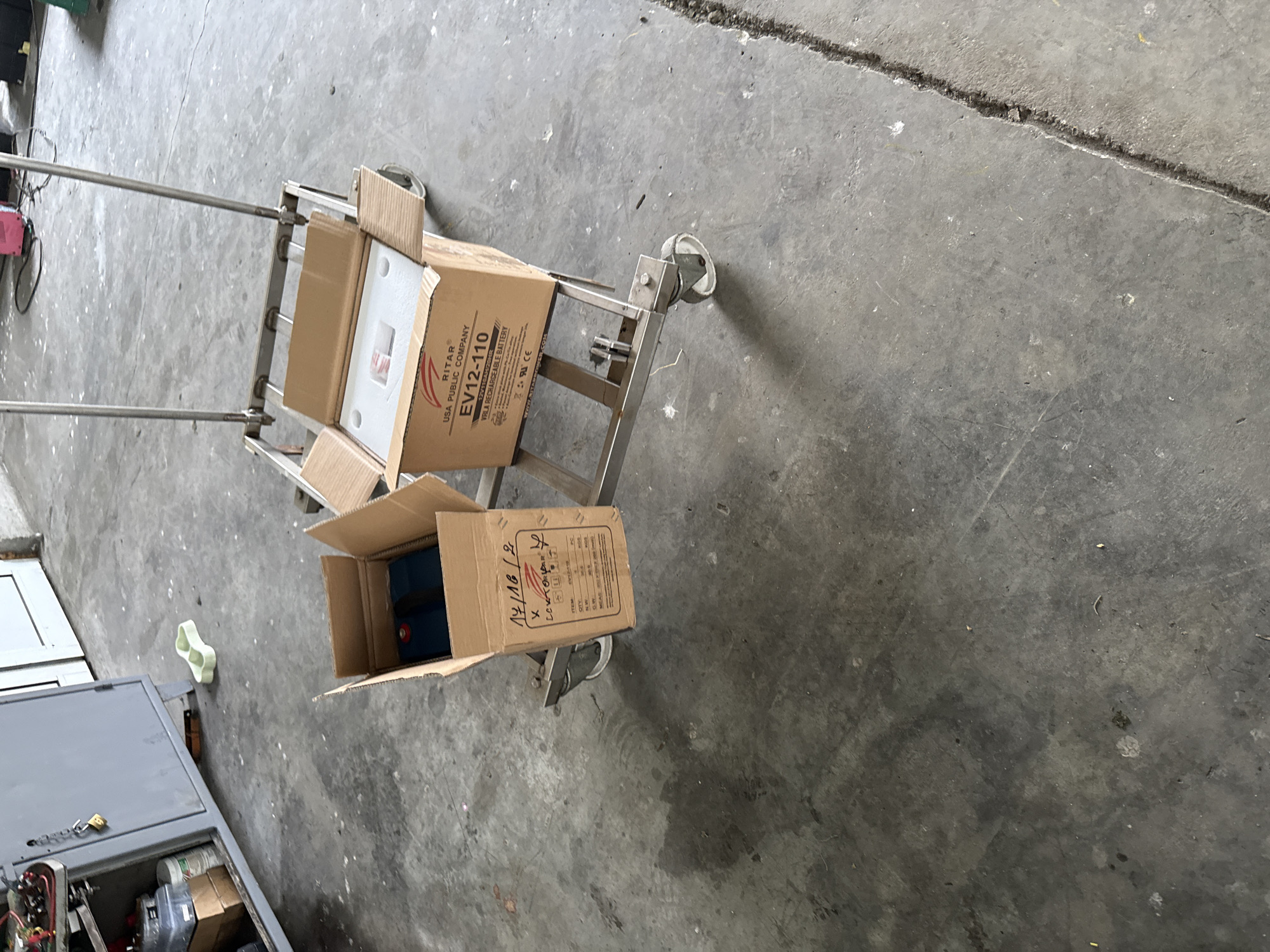 An overhead shot captures a metal hand truck or cart on a concrete floor, holding two open cardboard boxes. The scene appears to be an industrial or workshop environment.
The top box on the cart contains white foam packaging, likely protecting an item within. Visible text on this box includes "RITAR", "USA PUBLIC COMPANY", "EV12-110", "12V110AH/20HR", and "VRLA RECHARGEABLE BATTERY". It also states "GW: 33.5KG" and "NW: 30.5KG", indicating the gross and net weight.
The bottom box on the cart contains a blue object, which is likely another battery. Text on this box also prominently displays "RITAR" and "EV12-110", alongside "14/16/2", "LOKTORLAR", "Q'TY: 1 PC", "GW: 33.5KG", and "NW: 30.5KG".
The concrete floor shows wear and tear, with some cracks and debris. To the lower left, part of a grey electrical cabinet or machinery box is visible, with its lid open, revealing internal components and wires. A light green, bone-shaped object rests on the floor near the cabinet. No human activity is depicted. The lighting suggests it is daytime.