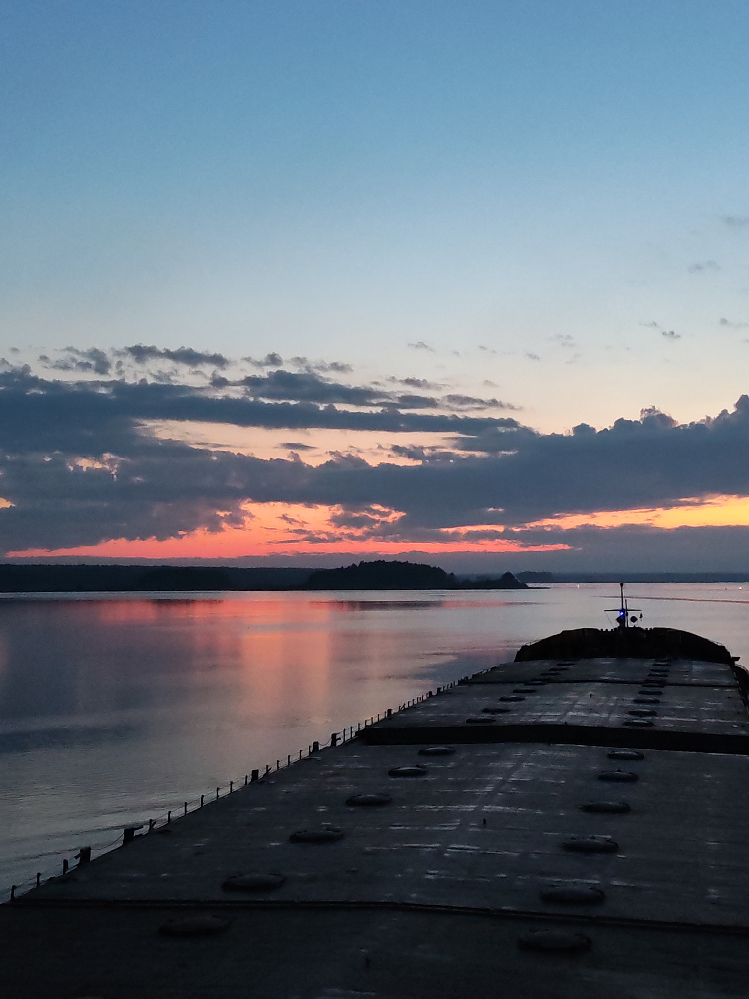 The image depicts a twilight scene over water, possibly a large lake or bay. The foreground is dominated by the dark, textured deck of a large ship or barge, running from the bottom of the frame and receding into the distance. The deck has a repetitive pattern of dark, oval shapes, likely hatches or access points.
Beyond the ship, the water reflects the colors of the sunset. The horizon line is defined by a dark band of land, possibly a forested area. Above the land, the sky displays a gradient of colors, transitioning from deep blue at the top to shades of orange, pink, and red near the horizon, where the sun is setting. Several layers of clouds are present, ranging in color from dark gray to lighter, pastel shades.
The overall lighting is dim, typical of twilight. The image does not contain any visible people. There is nothing in the image that indicates a location or association with Irdomatka, Russia.