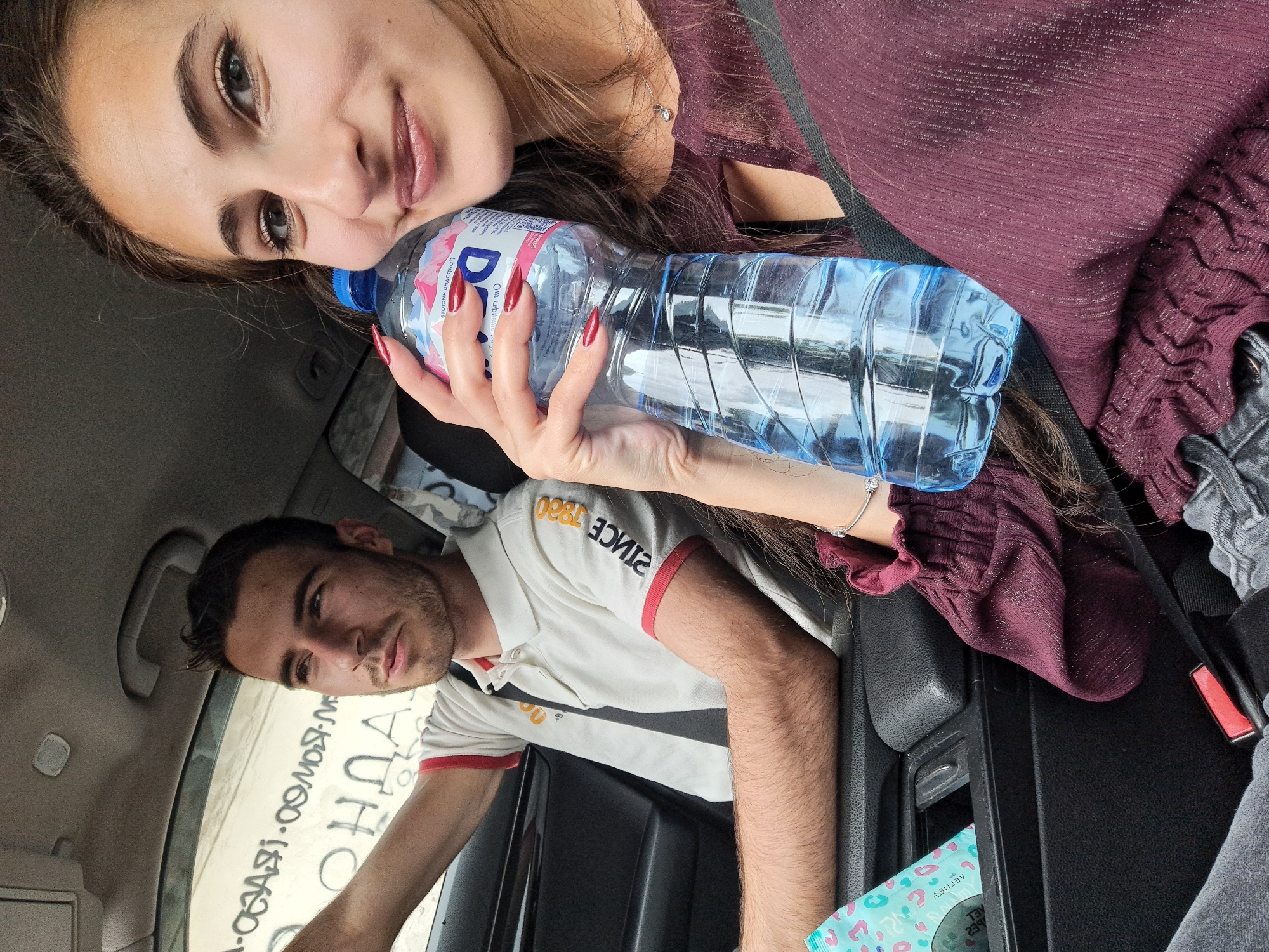 A selfie shows a woman and a man inside a vehicle. The woman, positioned at the top of the frame, holds a bottle of water with a blue cap. She has long brown hair and is wearing a maroon-colored top. The man is seated lower in the frame, wearing a white polo shirt with red accents. He has dark hair and a stubbly beard. The car's interior is visible around them, including a black center console with a pack of tissues inside and a safety belt. The setting appears to be indoors, potentially a car. The location of the city is unknown, with the city being unknown and state unknown.