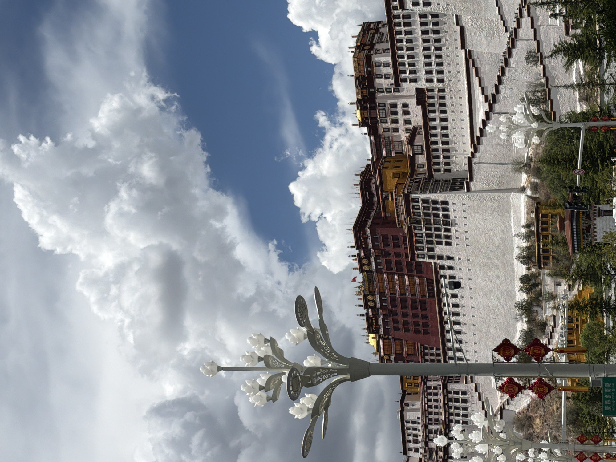 The image displays a majestic daytime view of the Potala Palace in Chengguan District, China, under a bright, partly cloudy sky. The imposing palace, built on a hill, is the central subject, showcasing its iconic red and white sections with numerous small, dark windows. Glimmers of golden roofs are visible on the upper structures of the palace.
In the foreground, a tall, decorative street lamp adorned with white, flower-like lights and intricate metalwork extends upwards from the left. Further to the right, red ornamental lanterns hang from another pole, adding vibrant cultural detail. Green foliage and trees are scattered around the base of the palace and along the hill, providing a natural contrast to the architecture.
The sky above is a clear blue, interspersed with large, fluffy white and grey clouds. A green street sign is partially visible, displaying Chinese characters that read "琼瑶多岗路" (Qiongyao Duogang Road), indicating a specific location. A traffic sign, possibly denoting a restriction, is also partly visible. The scene captures the grandeur of the historic site on what appears to be a pleasant, sunny day. No people are discernible in the frame, emphasizing the monumental scale of the palace.