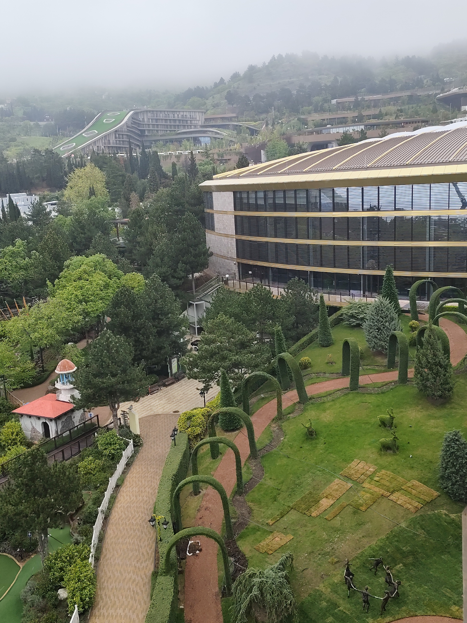 The image depicts an elevated view of a landscaped area within Симеизский поселковый совет, Ukraine. A modern building with a golden-colored roof and multiple rows of windows dominates the right side of the frame. Below, a meticulously maintained garden features shaped hedges forming archways over a path, topiary animals, and rectangular patches of exposed earth suggesting recent landscaping work. A circle of human-like sculptures is also visible on the grass. To the left, a small building with a red roof and white walls can be seen amidst dense greenery, suggesting a miniature village or decorative structure. In the background, another modern building with a grass-covered roof is partially obscured by fog or low clouds on the hills. The overall scene suggests a resort or upscale property in a mountainous region. The weather appears overcast.