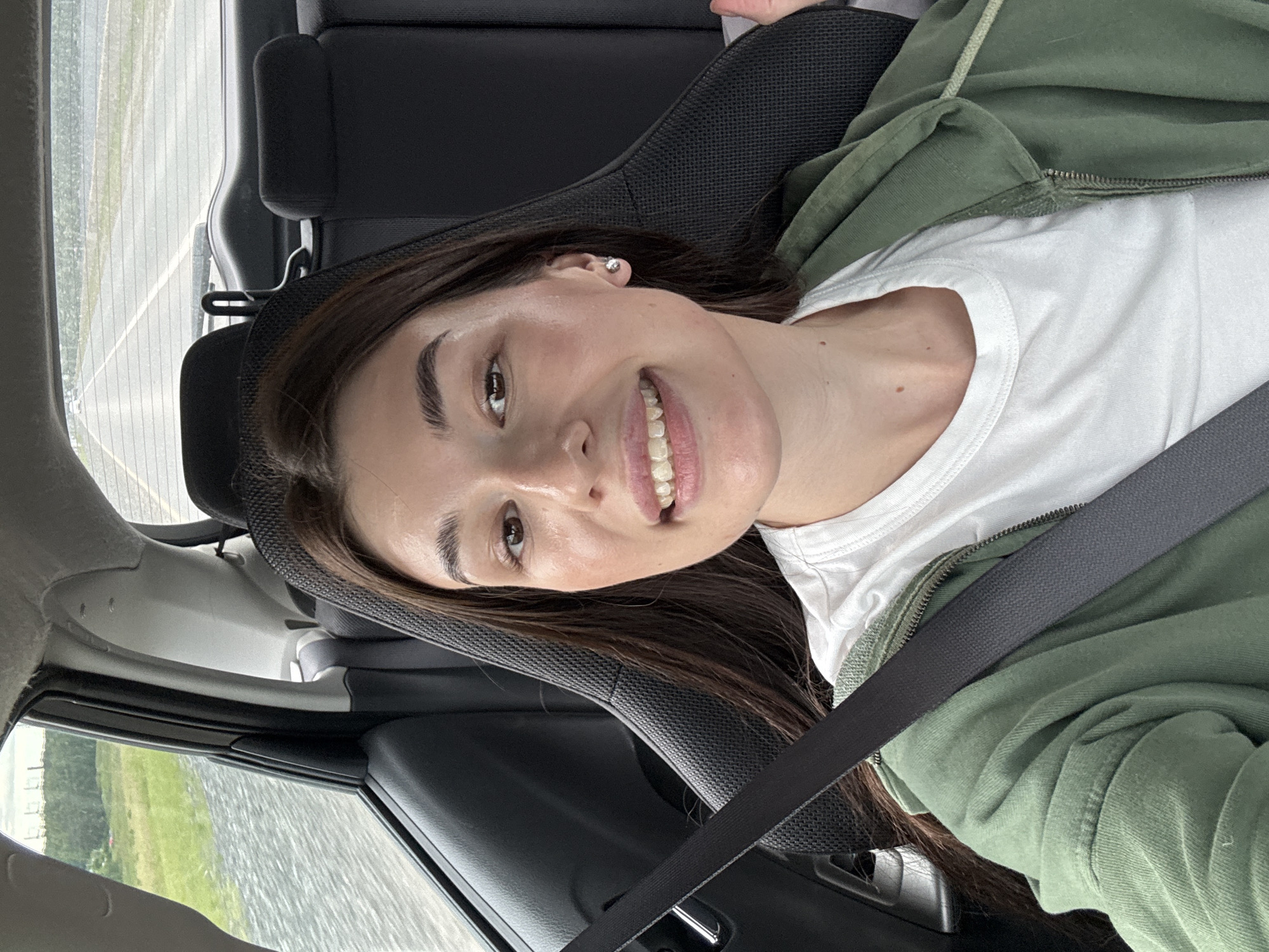The image captures a young woman seated in the front passenger seat of a car, smiling brightly towards the camera. She has long, dark brown hair and is wearing a white t-shirt layered under an open olive green hooded jacket or sweatshirt. A dark grey seatbelt diagonally crosses her chest. A small, round stud earring is visible in her right ear. Her expression is cheerful and content.
The setting is inside a vehicle, with parts of the car's dark interior, including a headrest and door panel, visible around her. Outside the car window, a wide body of water, likely a river or a lake (consistent with the Votkinsk Reservoir near Chaikovsky, Russia), stretches into the distance, with visible ripples on its surface. Beyond the water, a green landscape with trees and some distant buildings or structures can be discerned. The scene is illuminated by natural daylight, suggesting it is daytime. No visible text is present.