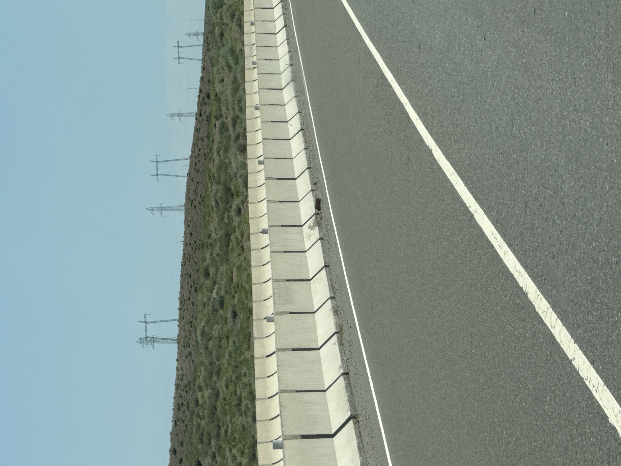 The image shows a road with concrete barriers on the left. Beyond the barriers, there is a grassy, hilly landscape with several tall electrical towers in the distance against a clear blue sky. The road has a white line marking its edge. The scene is likely an exterior shot of a highway or a major road. No people are visible in the image.